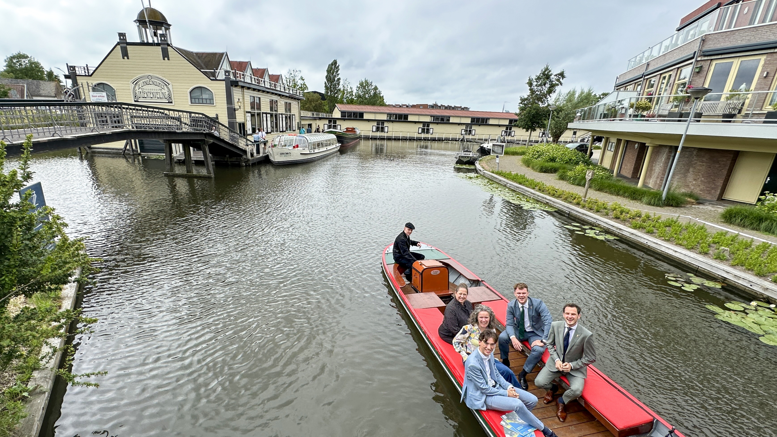 Een groep mensen zit in een rode rondvaartboot op een rustige waterweg naast historische gebouwen.