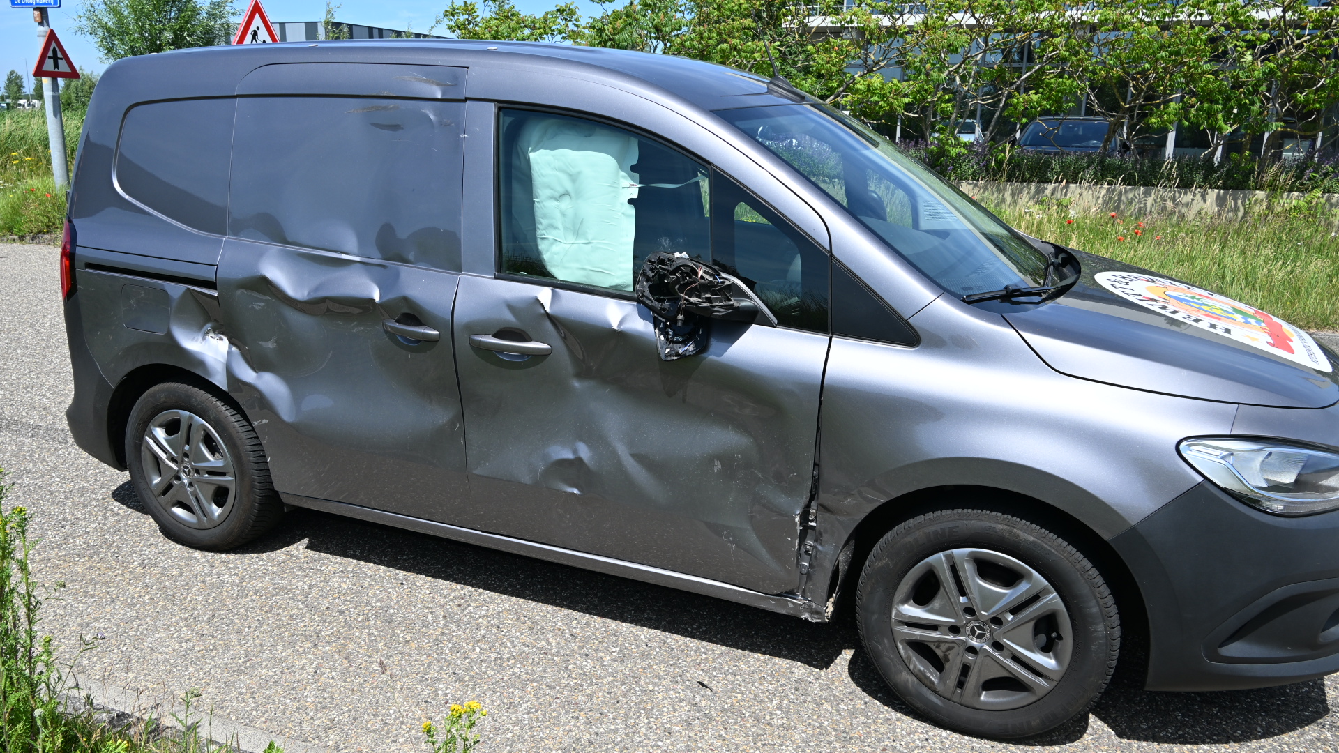 Grijze bestelwagen met aanzienlijke deuken aan de zijkant en een kapotte zijspiegel, geparkeerd op een weg.