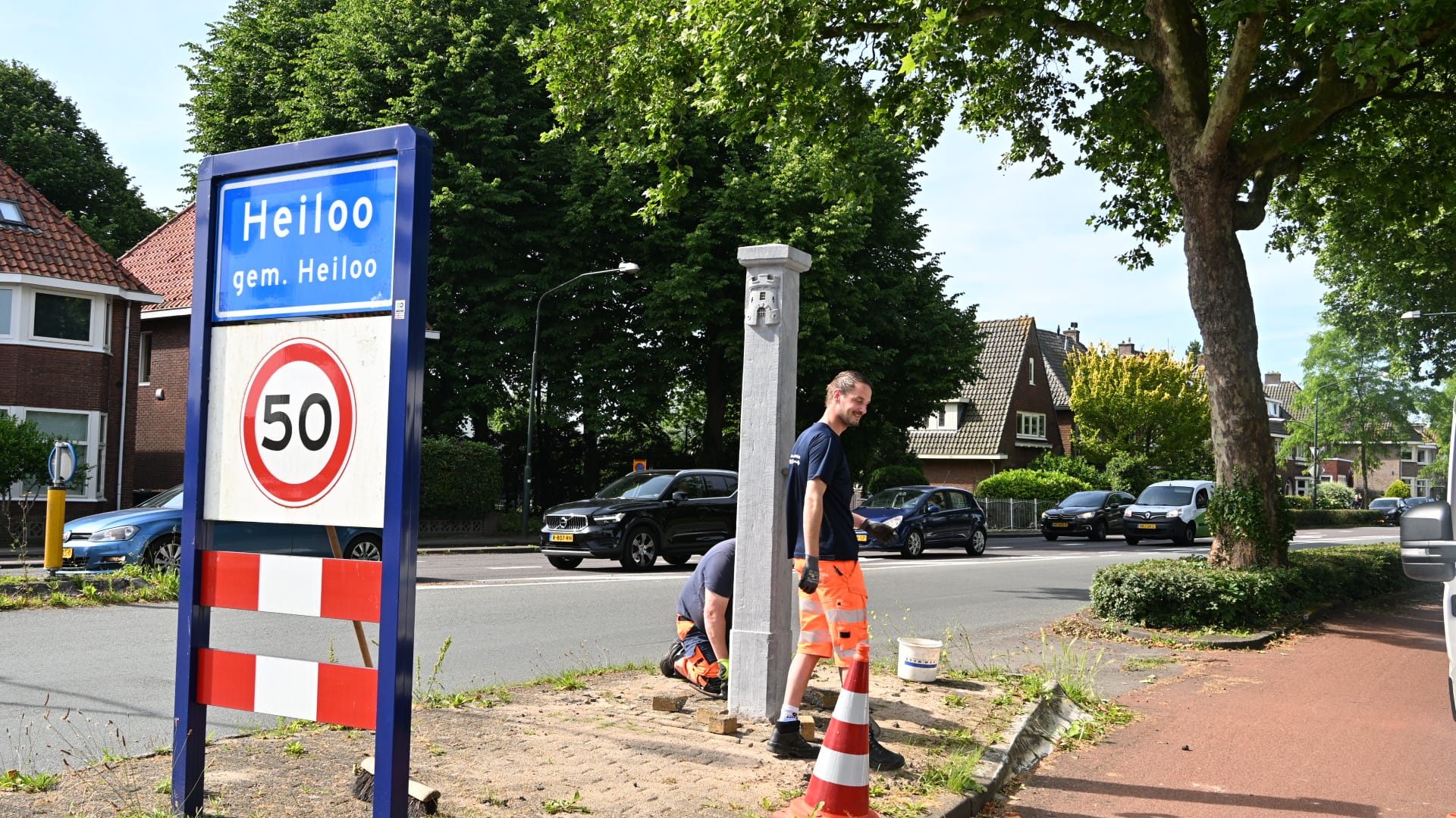 Verkeersbord met "Heiloo" en snelheidslimiet 50 km/u, naast een man die werkt aan een paal op een zonnige straat.