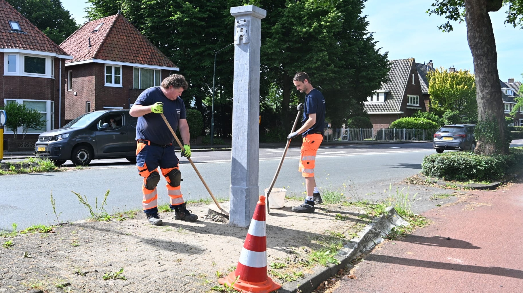 Twee mannen in werkkleding vegen rondom een betonnen paal op een stoep langs een straat, met een verkeerskegel erbij.