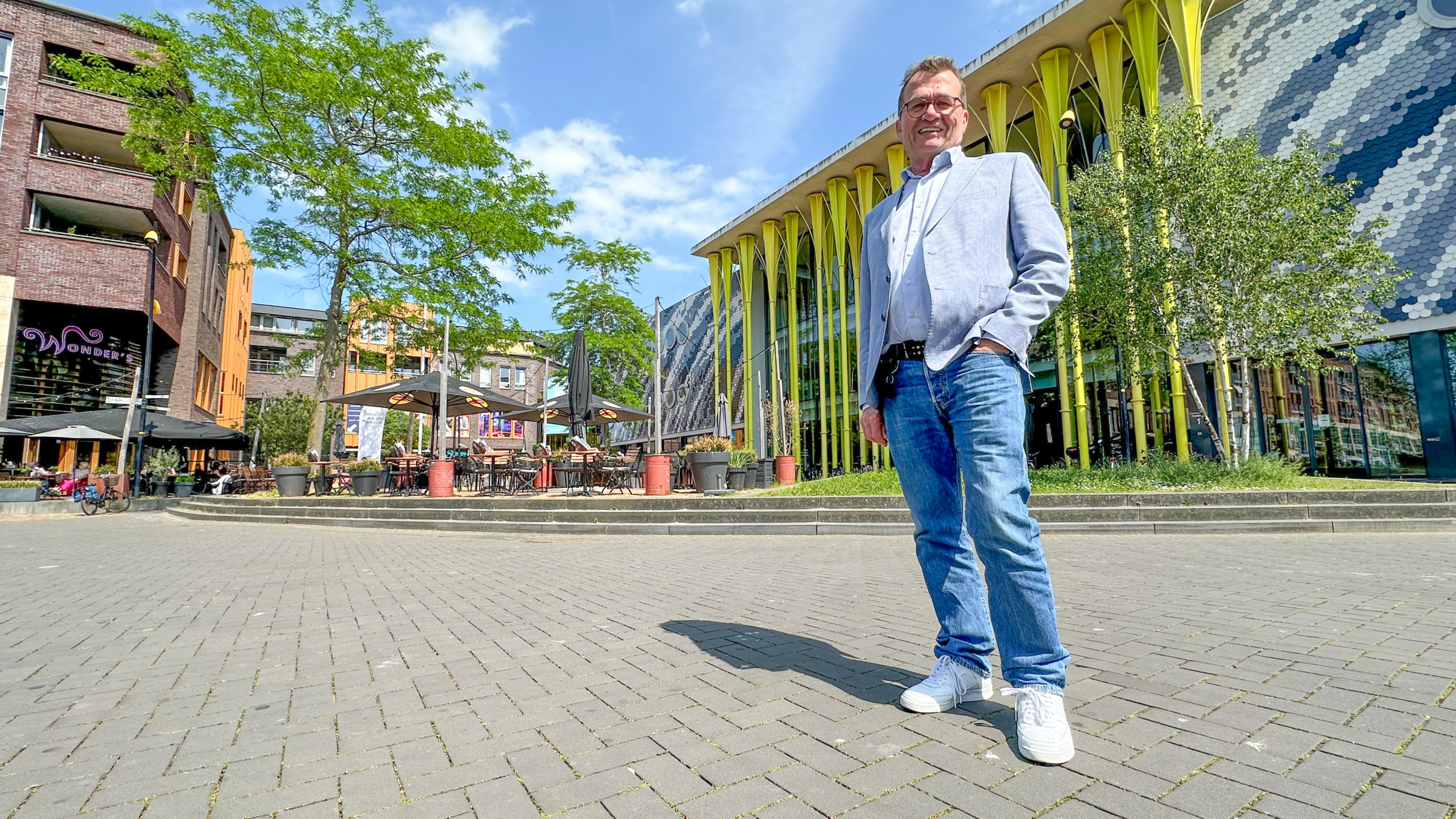 Man in casual outfit poseert voor modern gebouw met geelgroene pilaren en groot terras op een zonnige dag.
