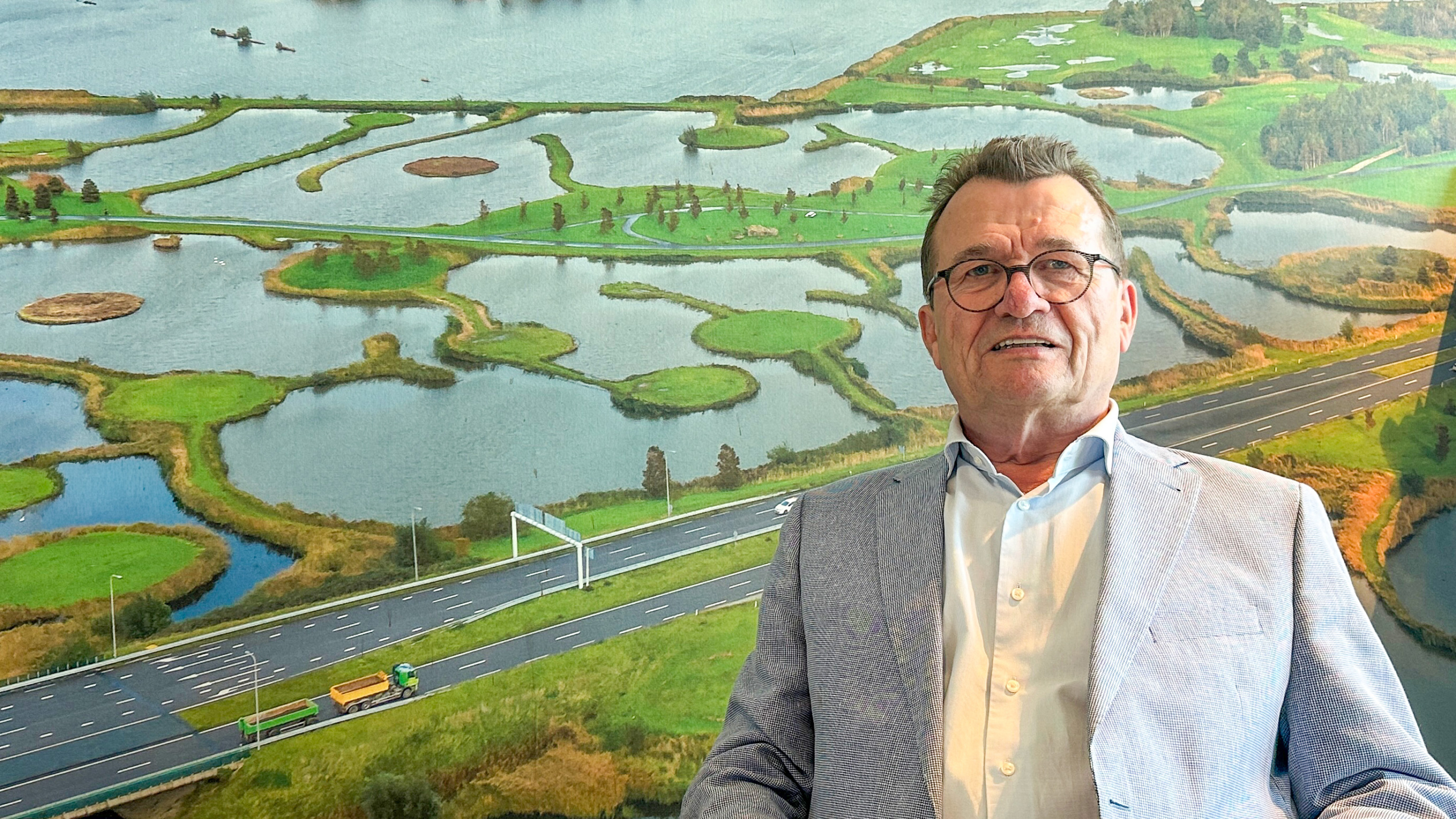 Man met bril en lichtblauw colbert staat voor een muur met een grote foto van een weg en een waterrijk landschap.