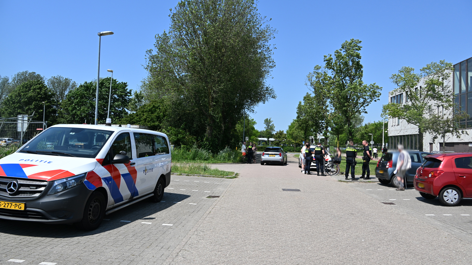 Politieauto en agenten op straat in gesprek met mensen, omgeven door bomen en een kantoorgebouw op de achtergrond.