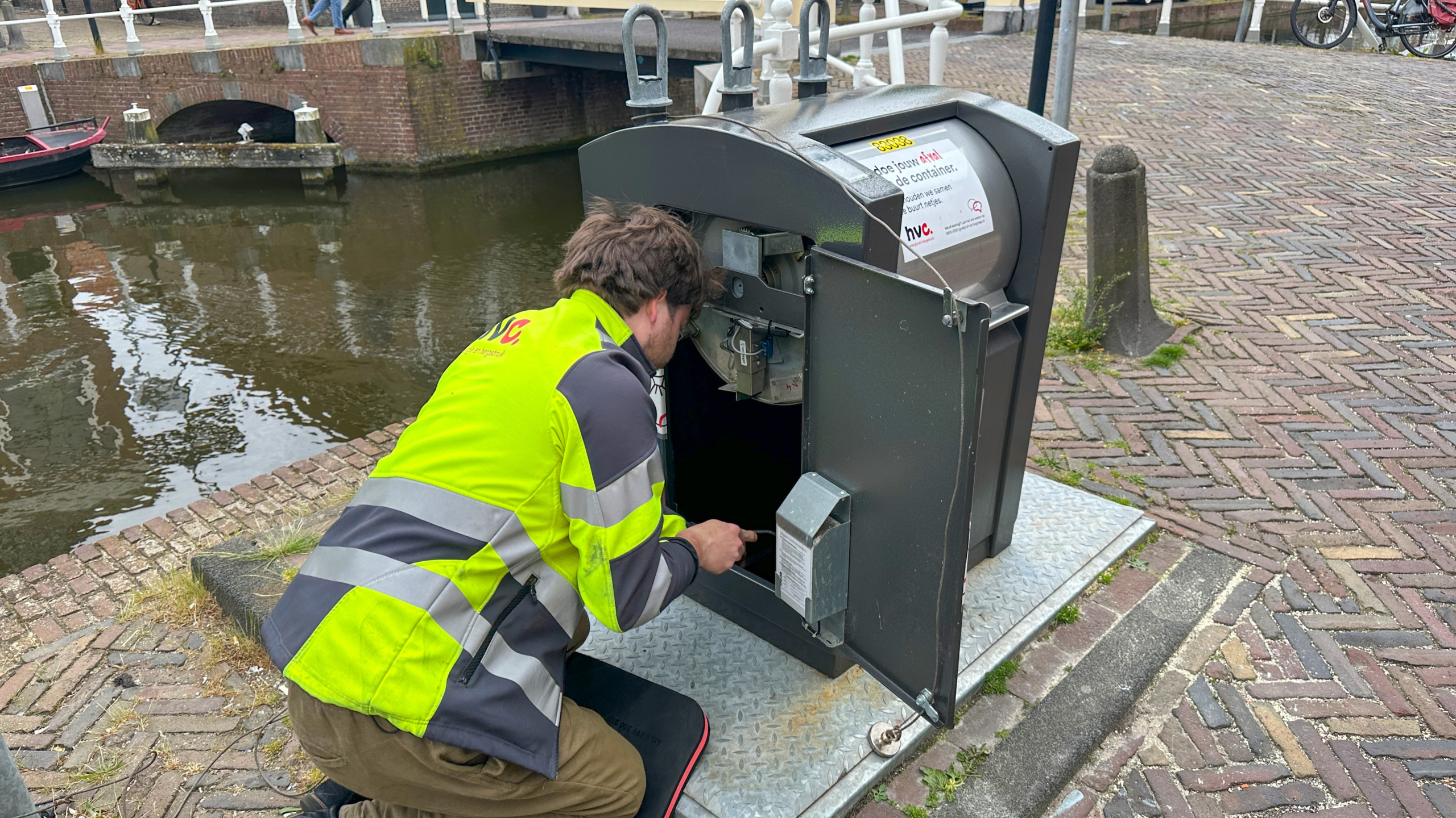 Een man in een geel veiligheidsvest werkt aan een ondergrondse container bij een gracht in een stedelijke omgeving.