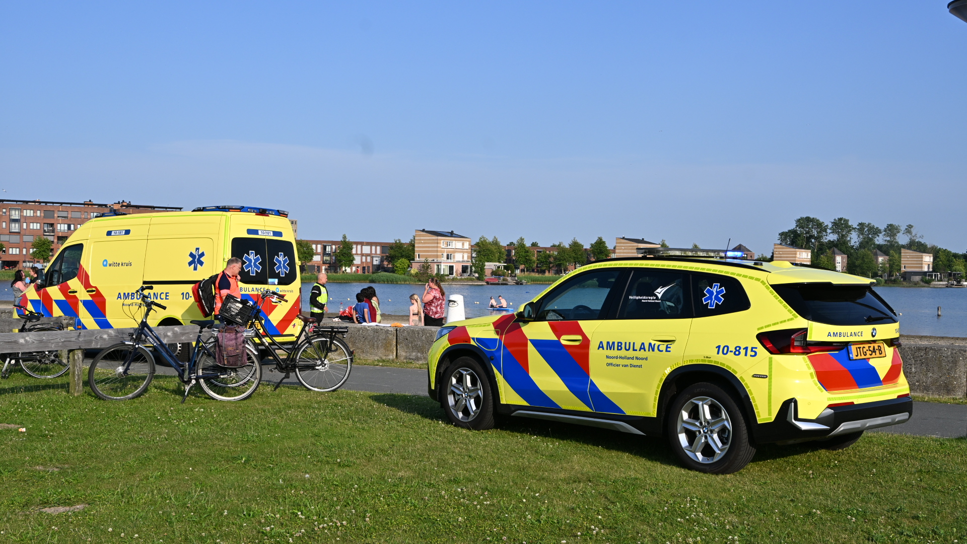 Twee ambulancevoertuigen staan geparkeerd bij een waterkant, omringd door enkele mensen en fietsen.