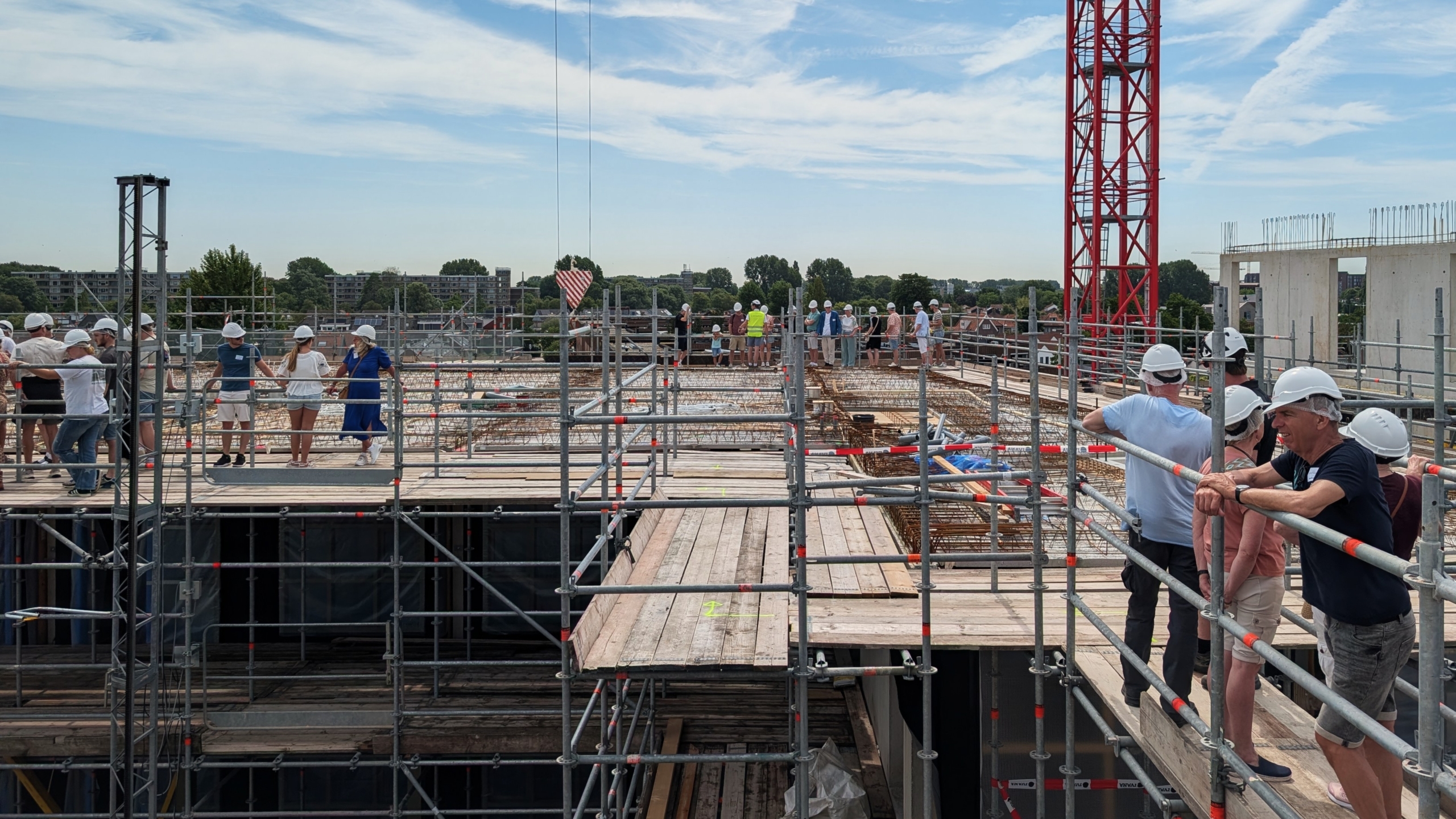 Mensen met helmen lopen rond op een constructieplaats met steigers en een kraan, onder een heldere lucht.