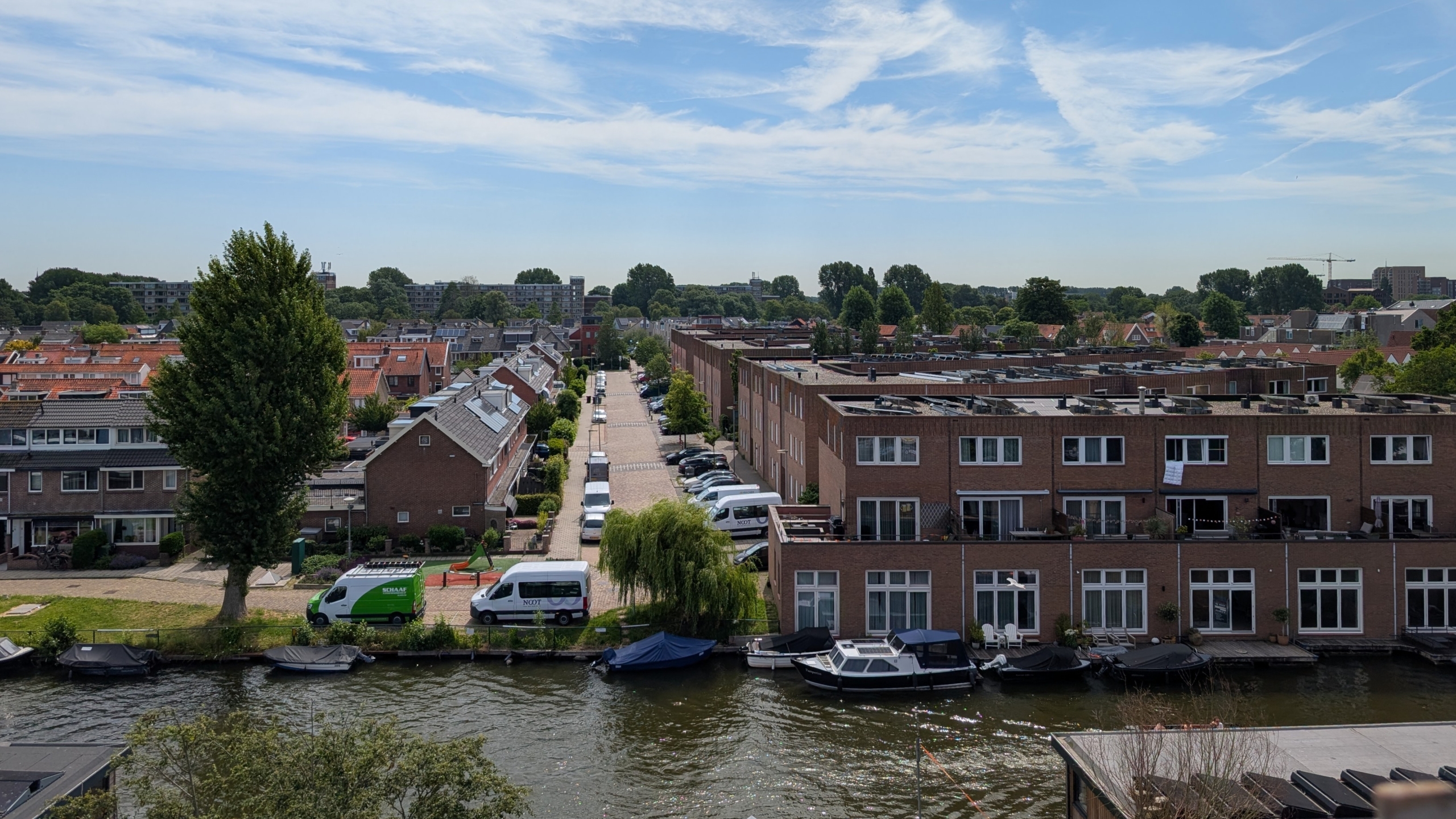 Bovenaanzicht van een rustige Nederlandse woonwijk langs een gracht, met rijen bakstenen huizen en geparkeerde auto's.