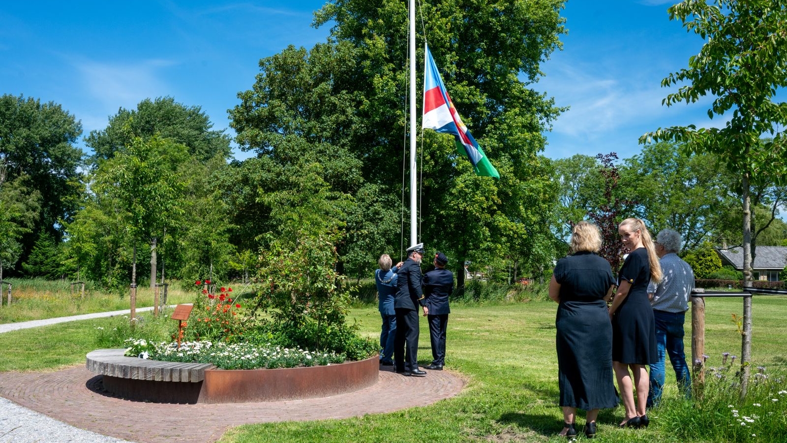 Mensen hijsen een vlag in een parkachtige omgeving, omgeven door bomen en een bloembed.