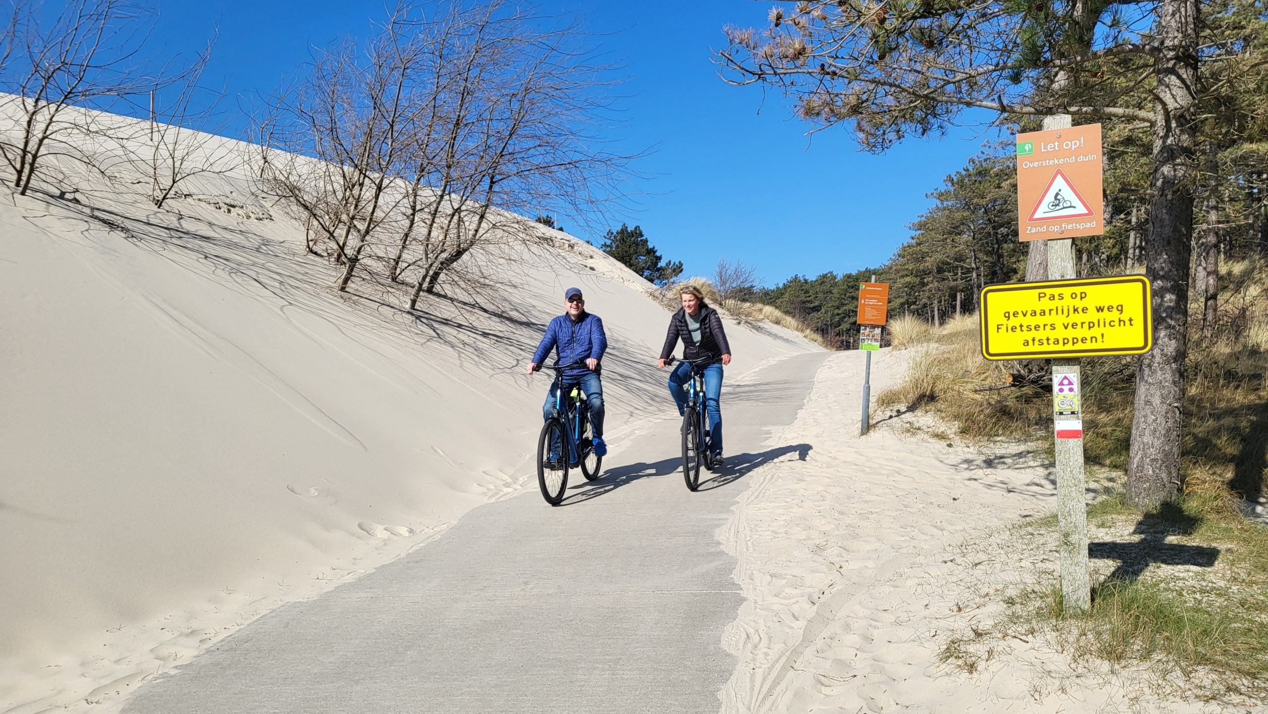 Twee mensen fietsen over een pad door duinen met waarschuwingsborden voor overstekend zand.