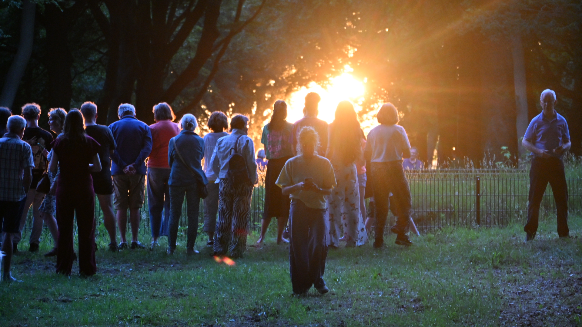 Een groep mensen staat in een park bij zonsondergang, met stralen van het licht door de bomen.