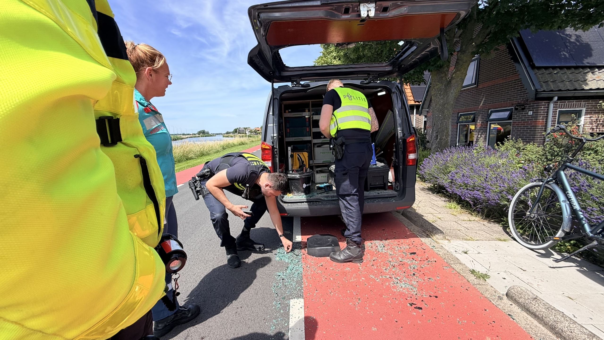 Politieagenten onderzoeken glasscherven op een fietspad naast een geopende bestelwagen, in een landelijke omgeving.