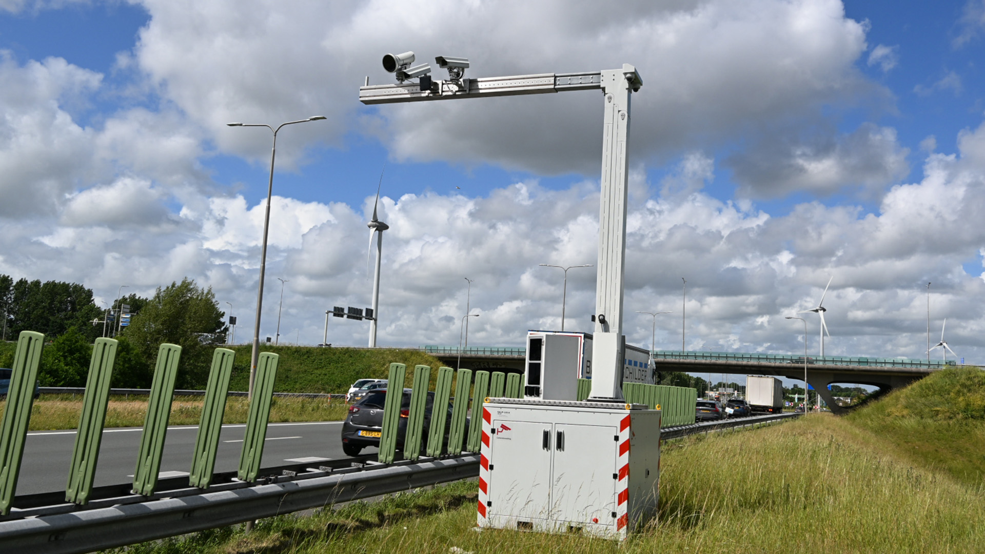 Verkeerscamera's op een paal langs een snelweg met auto's, windturbines en een brug op de achtergrond onder een bewolkte hemel.