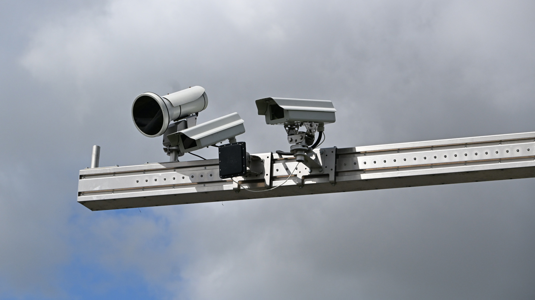 Drie verkeerscamera's gemonteerd op een metalen balk tegen een bewolkte lucht.