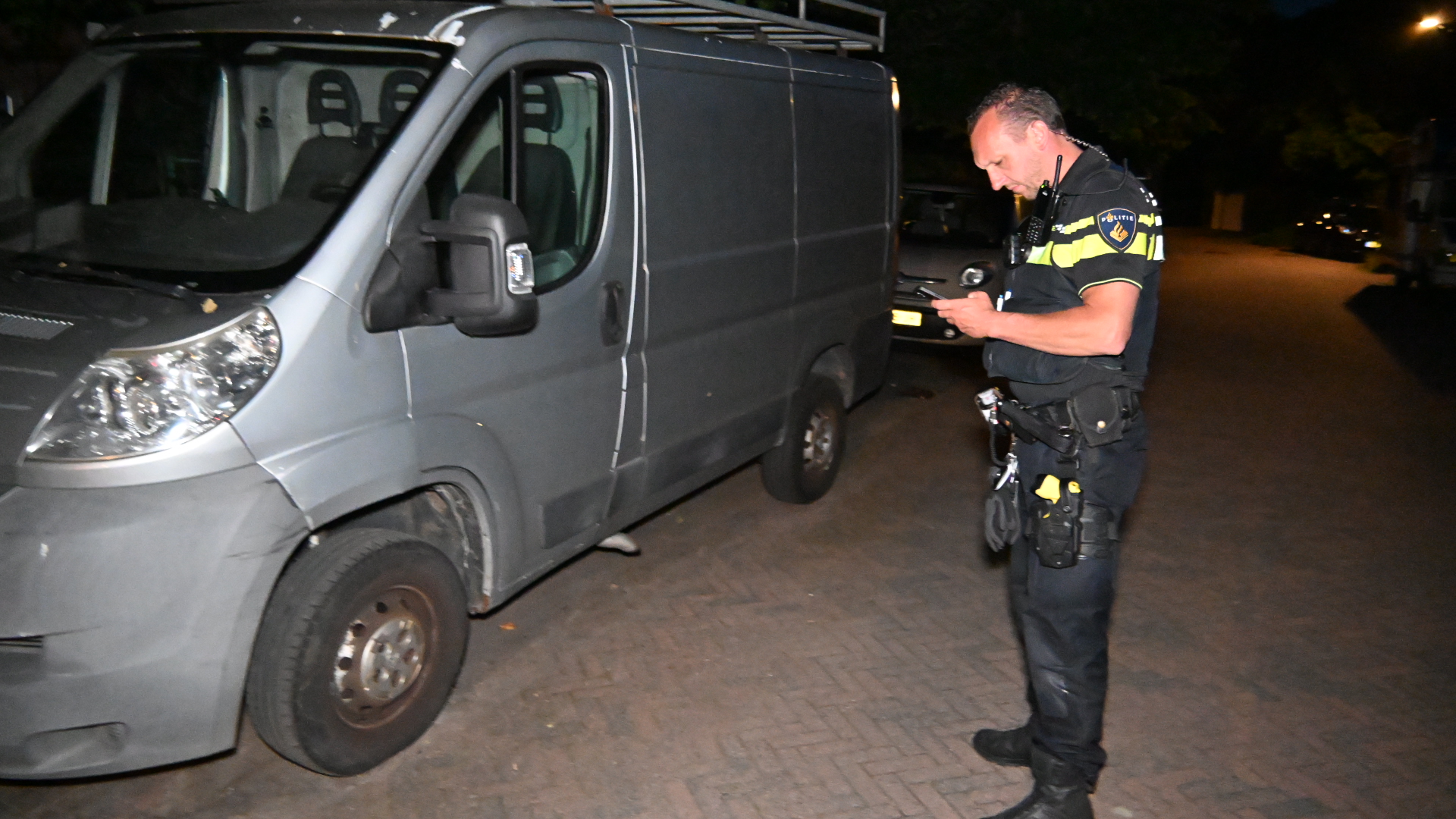 Een politieagent controleert zijn telefoon naast een geparkeerde grijze bestelbus op een straat in het donker.