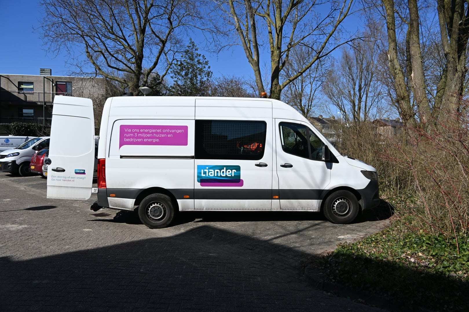 Witte bestelwagen van Liander geparkeerd bij bomen; deurschildering promoot energievoorziening voor miljoenen huizen.