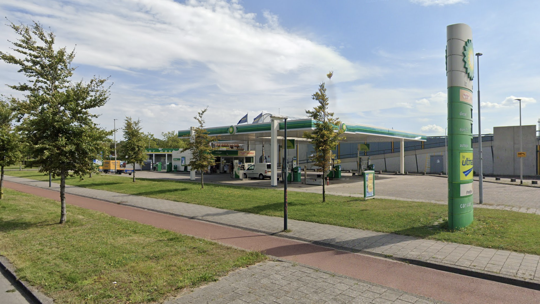 Tankstation langs een straat met een BP-bord en diverse bomen op de groenstrook onder een blauwe hemel.