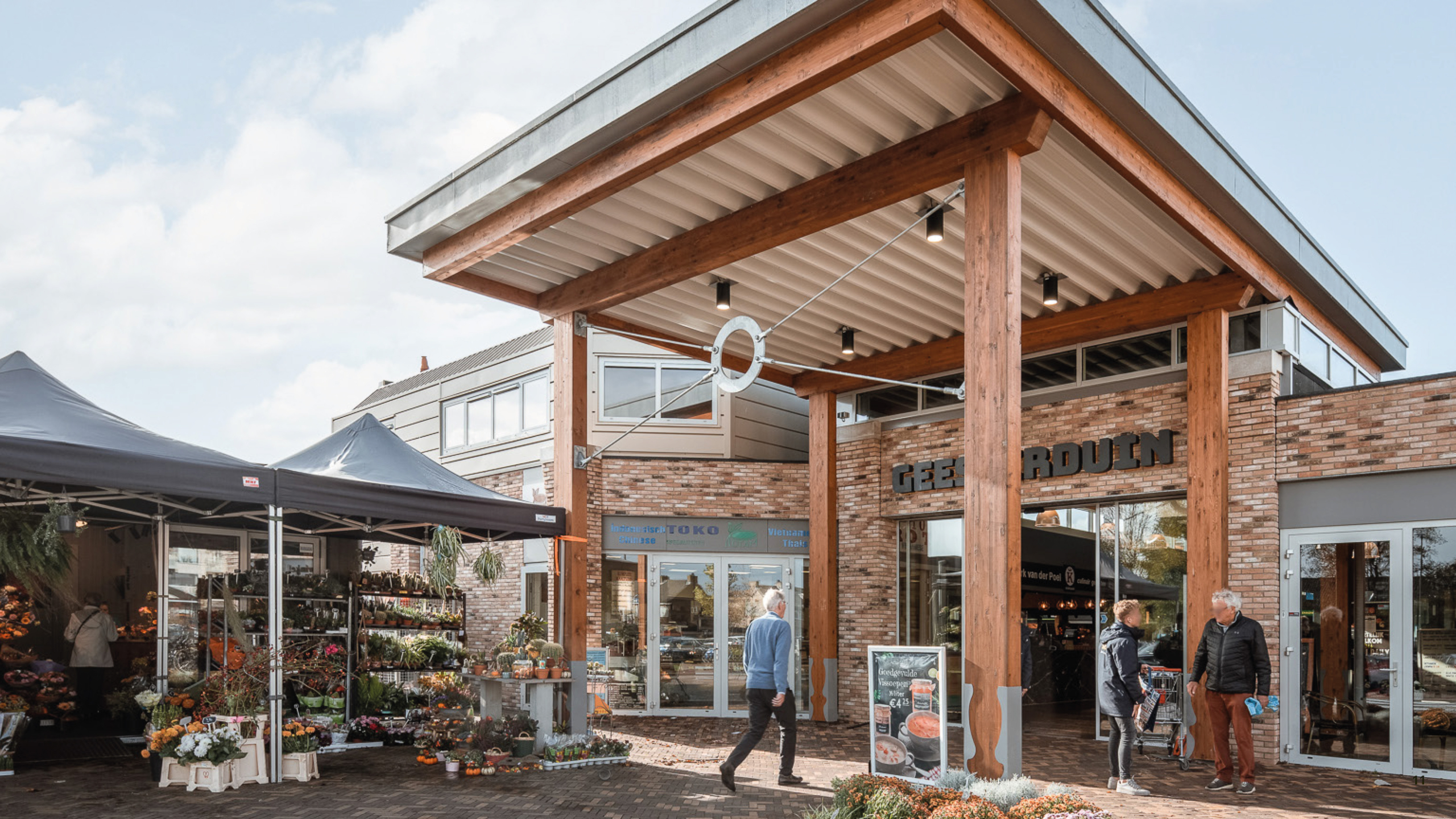 Ingang van een winkelcentrum met houten pilaren, kraam met planten en bloemen, en enkele mensen die rondlopen.