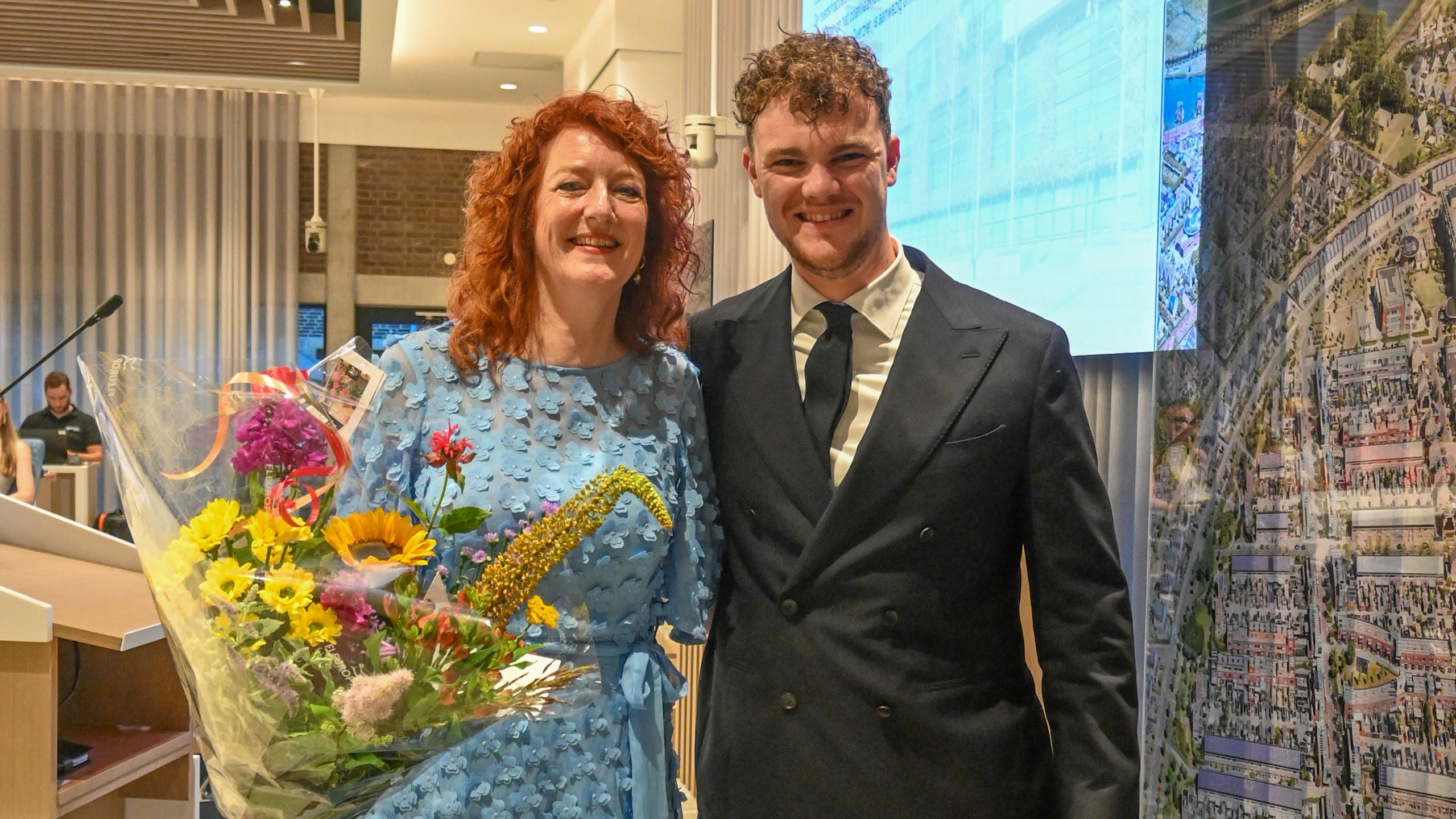 Een vrouw in een blauwe jurk met een boeket bloemen en een man in een donker pak staan samen te glimlachen.