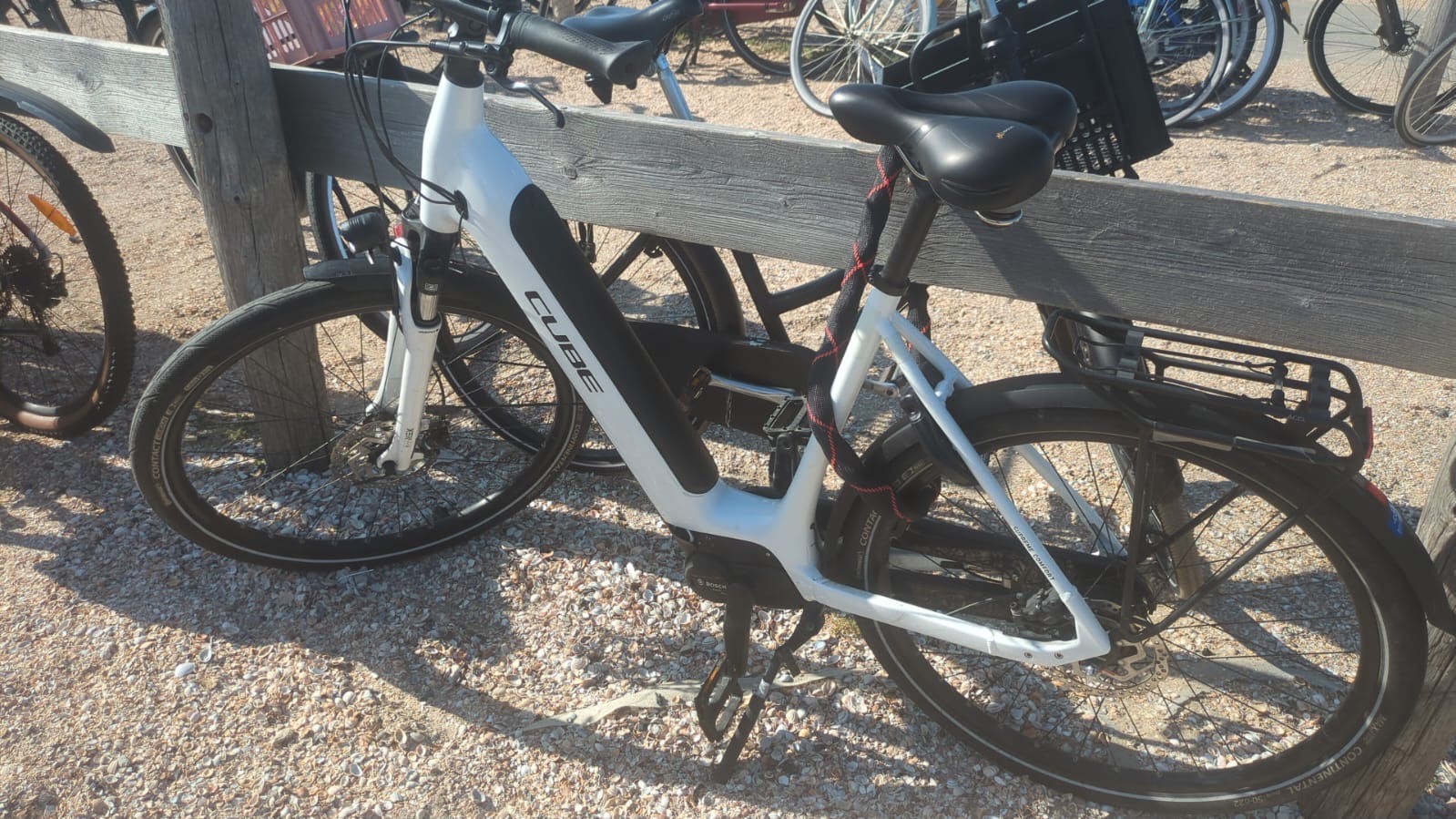 Witte elektrische fiets van het merk Cube geparkeerd tegen een houten hek op een grindoppervlak.