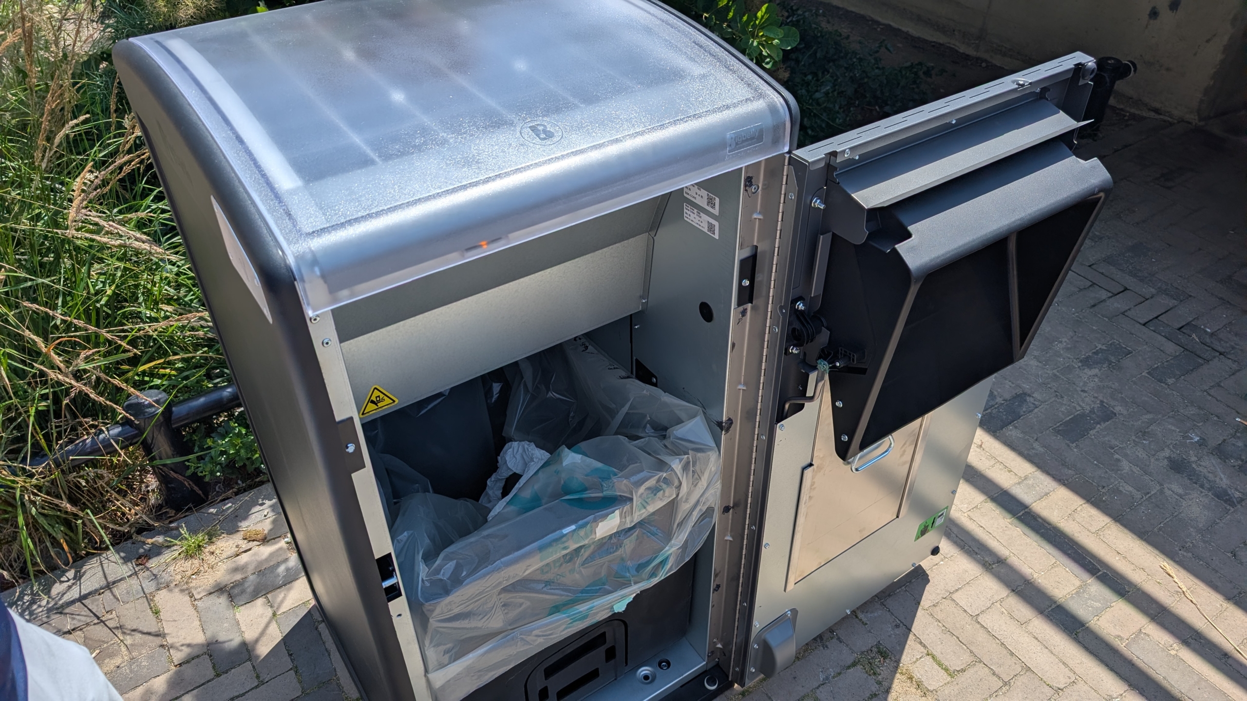 Openstaande ondergrondse afvalcontainer op een zonnige dag, met plastic zakken zichtbaar binnenin.