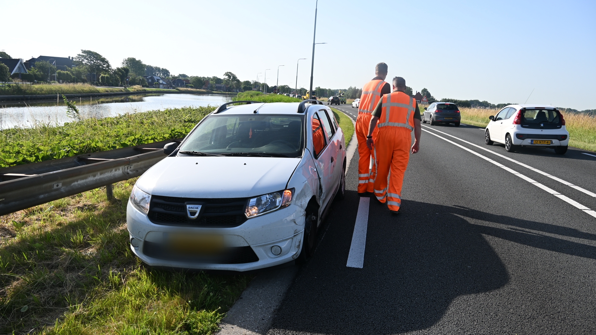 Beschadigde witte auto naast vangrail, met twee mannen in oranje werkkleding dichterbij op de weg.