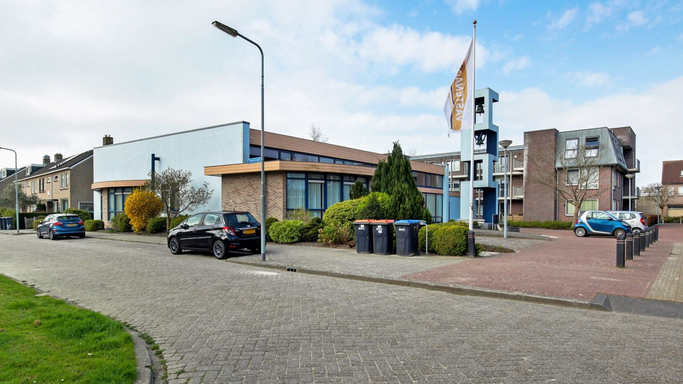 Straatbeeld met moderne gebouwen, geparkeerde auto's, struiken en een vlag met tekst "SWSH".