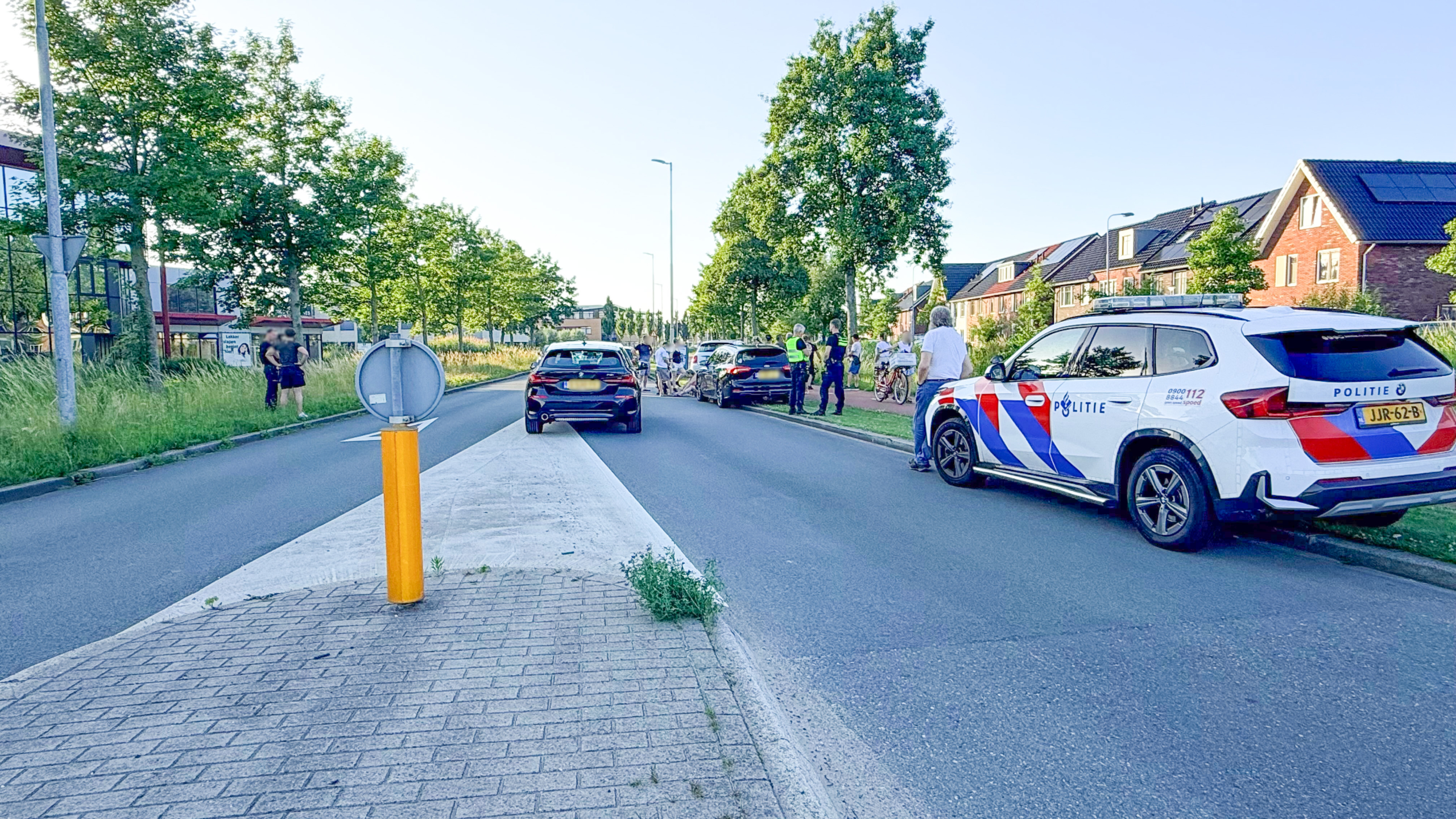 Politieauto en mensen langs de weg, met een auto op de rijbaan en fietsers aan de kant.
