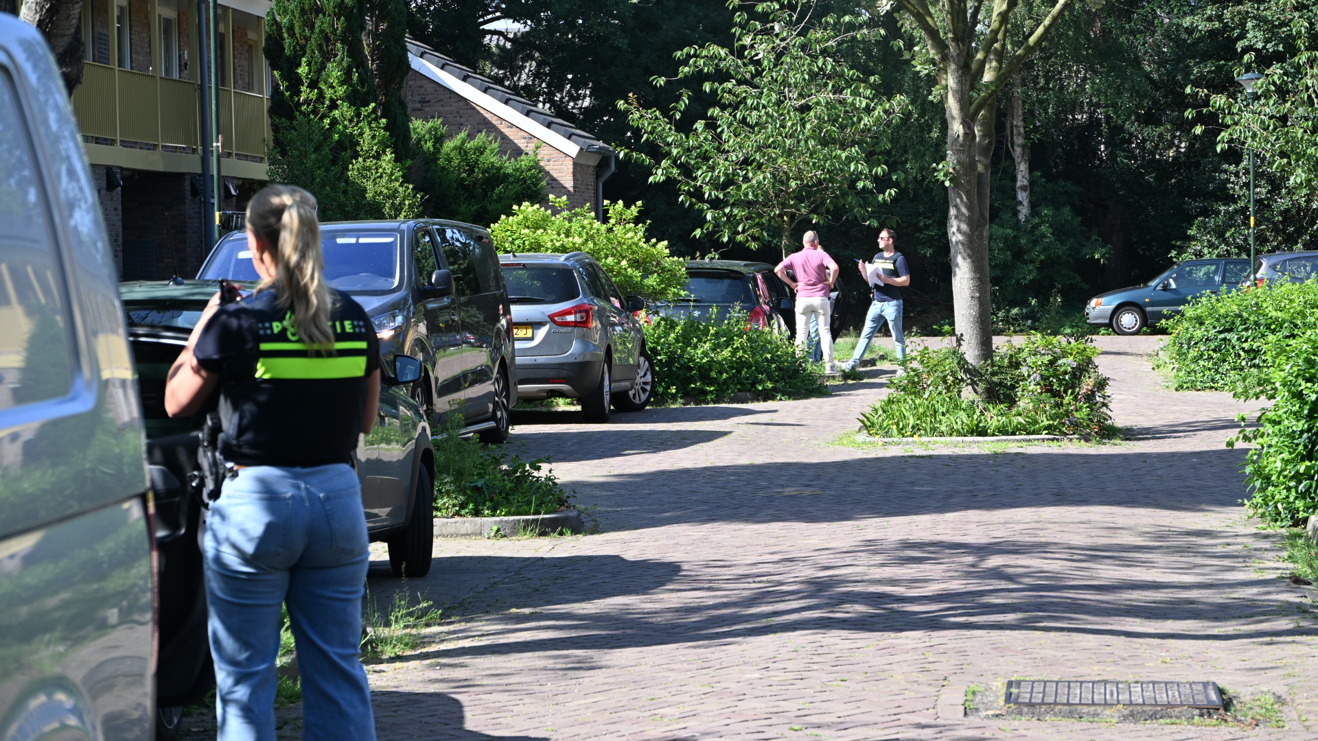 Een agent met politie-uniform observeert een parkeergelegenheid waar meerdere auto's staan en twee mannen een gesprek hebben.
