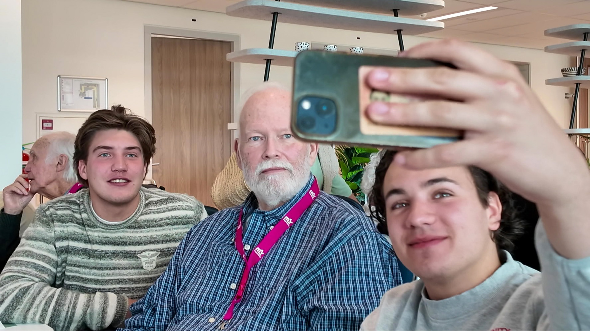 Twee jonge mannen en een oudere man maken een selfie in een binnenruimte.