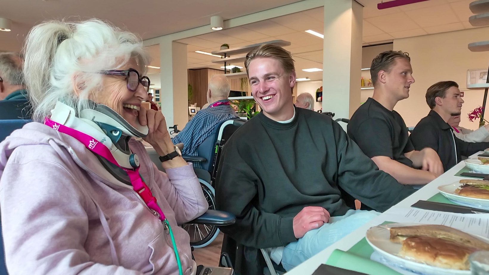 Een oudere vrouw met een nekkraag lacht terwijl ze met een jonge man praat aan een tafel met eten.