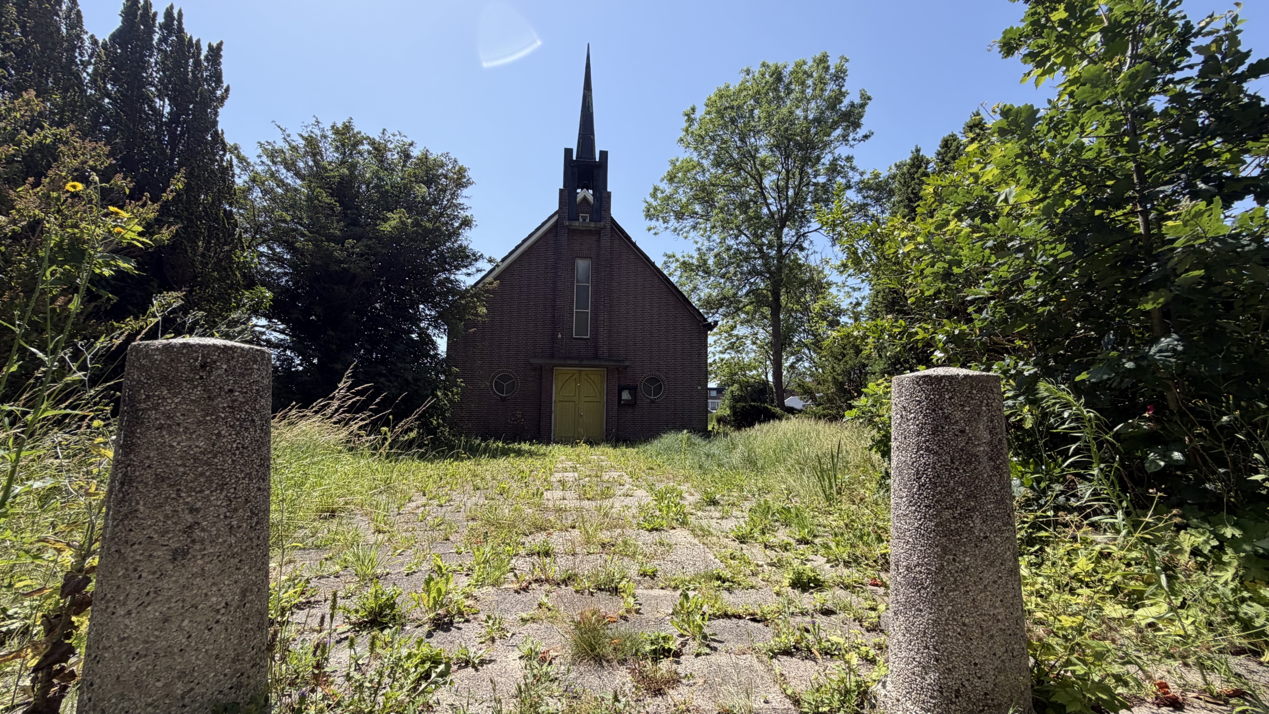 Kleine bakstenen kerk met torenspits, omgeven door bomen en gras, gezien vanaf een pad met betonnen palen.