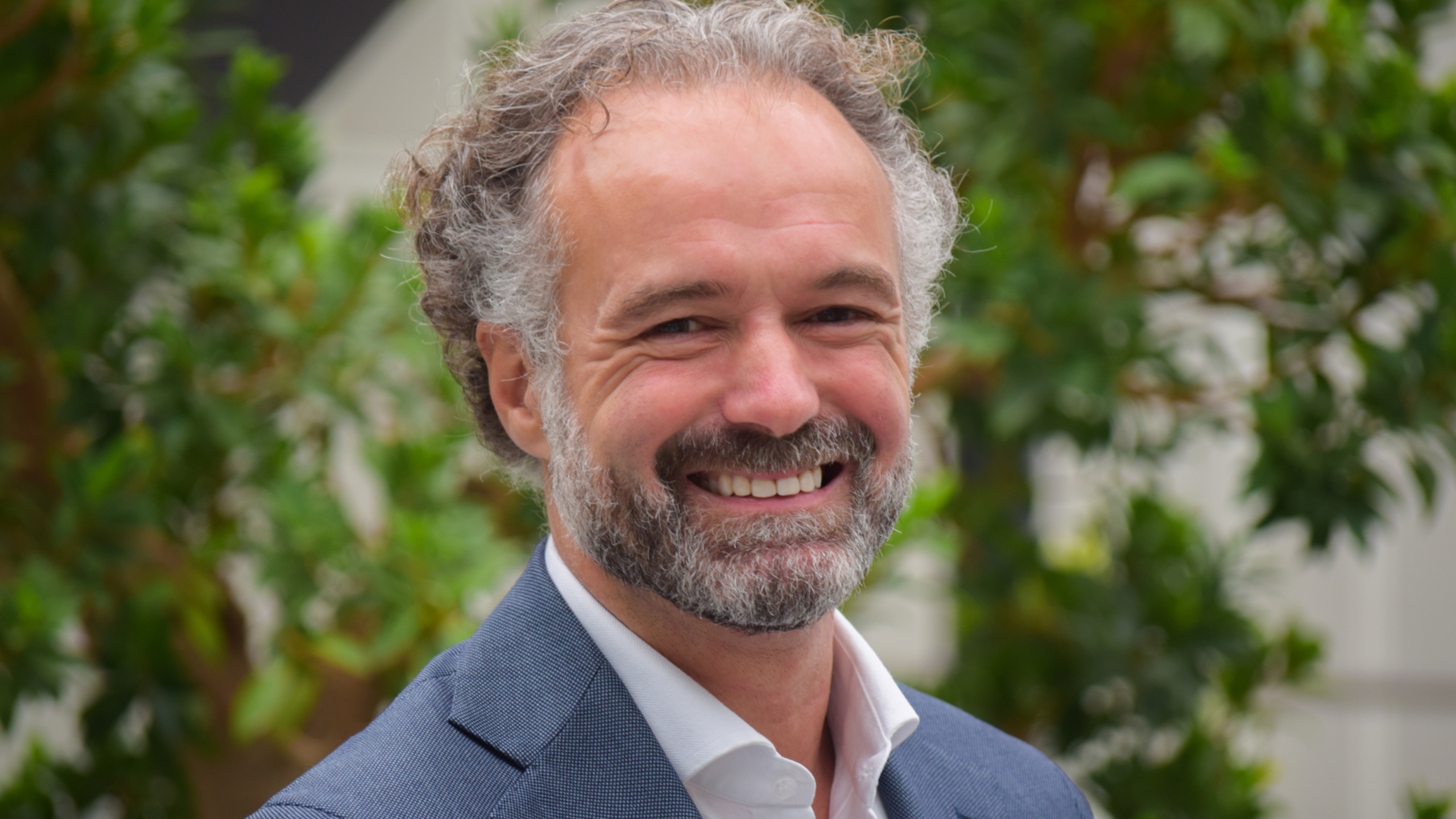 Lachende man met baard en krullend haar draagt een blauw colbert, met groene planten op de achtergrond.