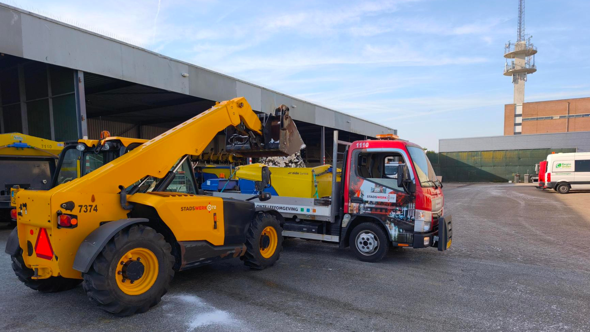 Een gele vorkheftruck laadt zout in een rode vrachtwagen op een open terrein met bedrijfspanden op de achtergrond.