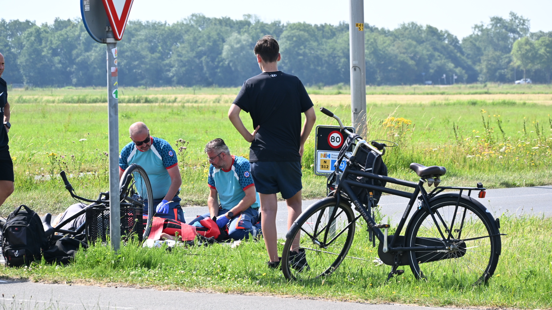 Twee mensen in sportkleding verlenen hulp aan een persoon naast een fiets, met een kijker en een verkeersbord op de achtergrond.