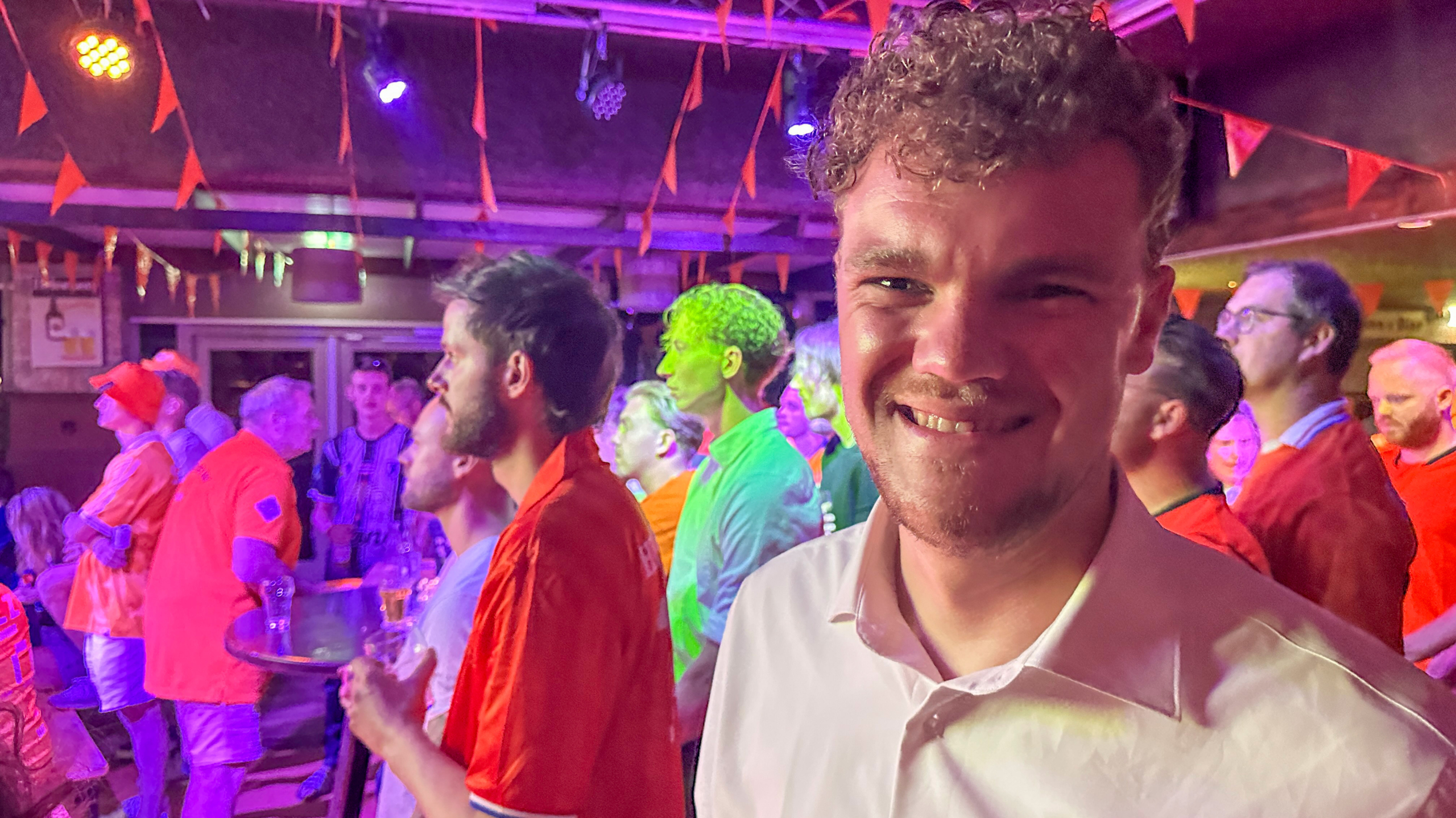 Man glimlacht in een feestelijke ruimte met oranje versieringen en mensen die een scherm volgen.