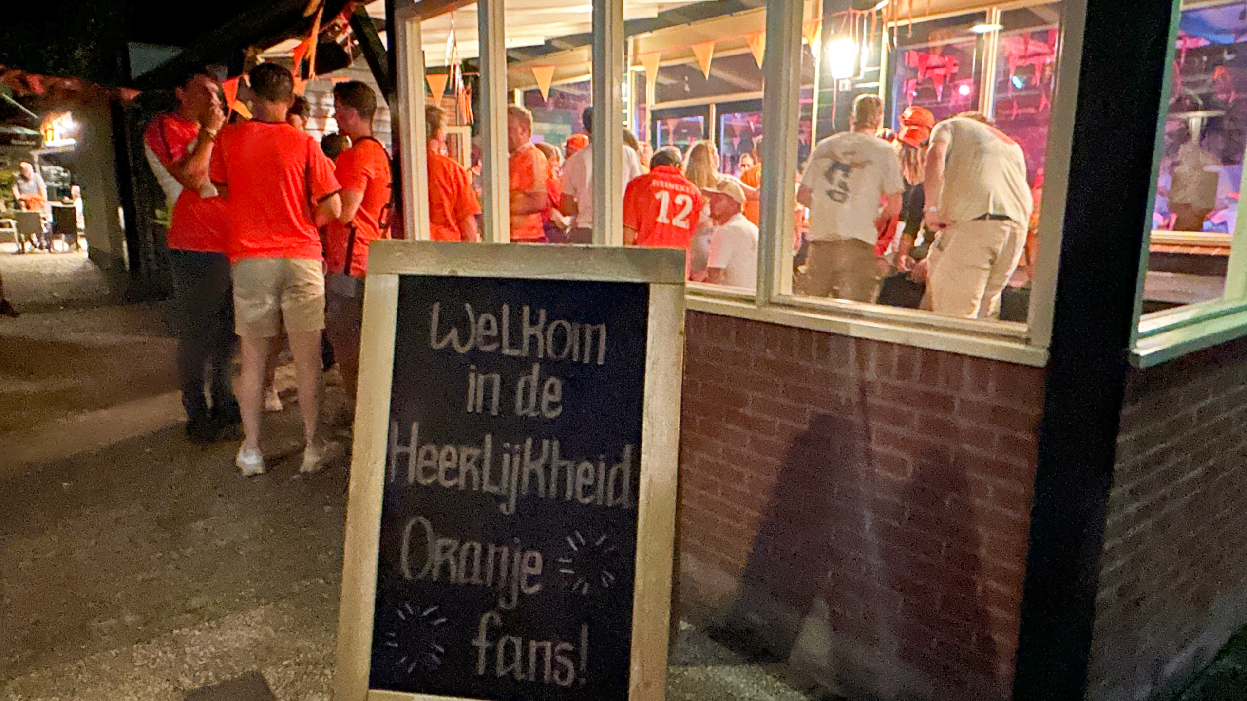 Groep mensen in oranje shirts staat bij een café met een bord met de tekst "Welkom in de Heerlijkheid Oranje fans!"