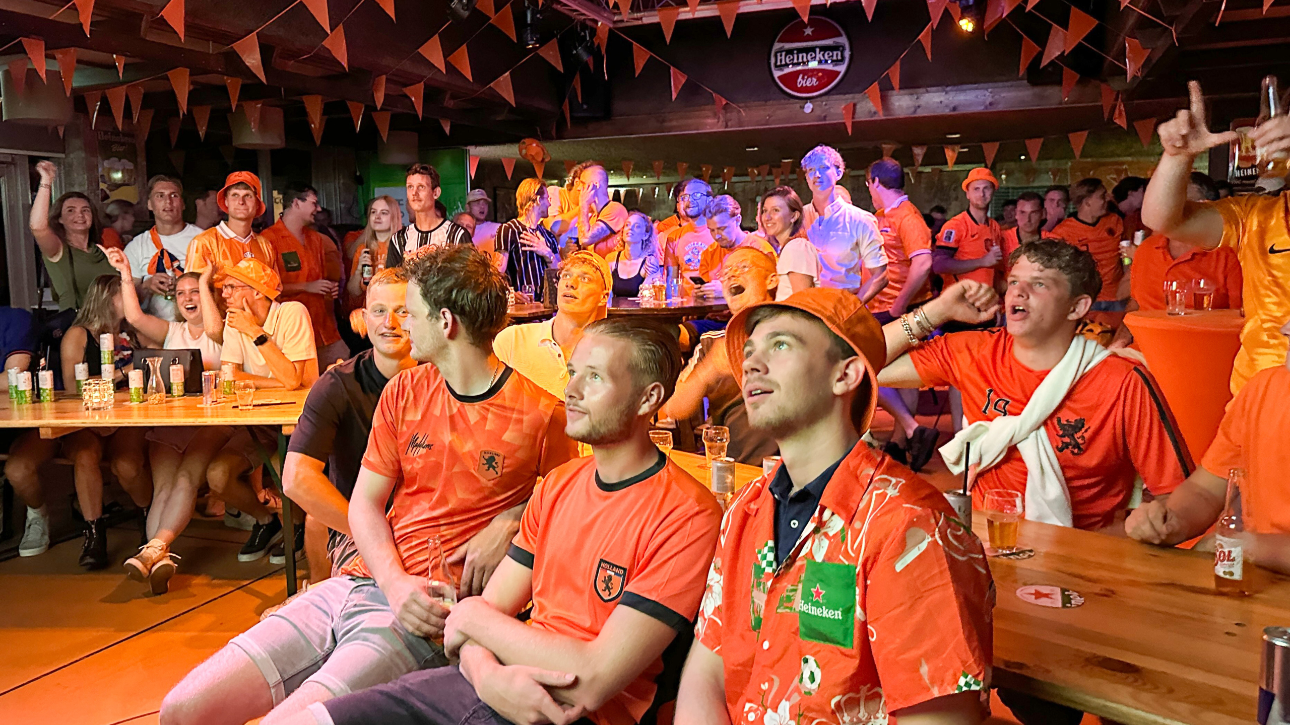 Mensen in een kroeg, gekleed in oranje, kijken aandachtig naar een scherm, vermoedelijk voor een sportwedstrijd.