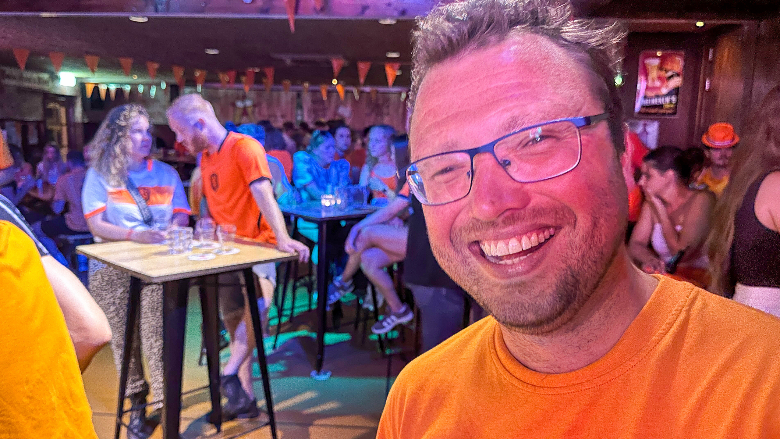 Man met bril glimlacht in een bar vol mensen gekleed in oranje, waarschijnlijk tijdens een evenement of wedstrijd.