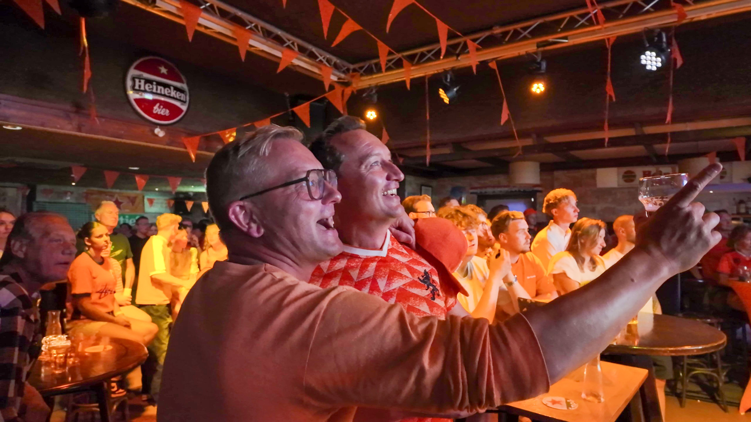 Mensen kijken naar een scherm in een café versierd met oranje vlaggetjes; een man houdt een glas omhoog.