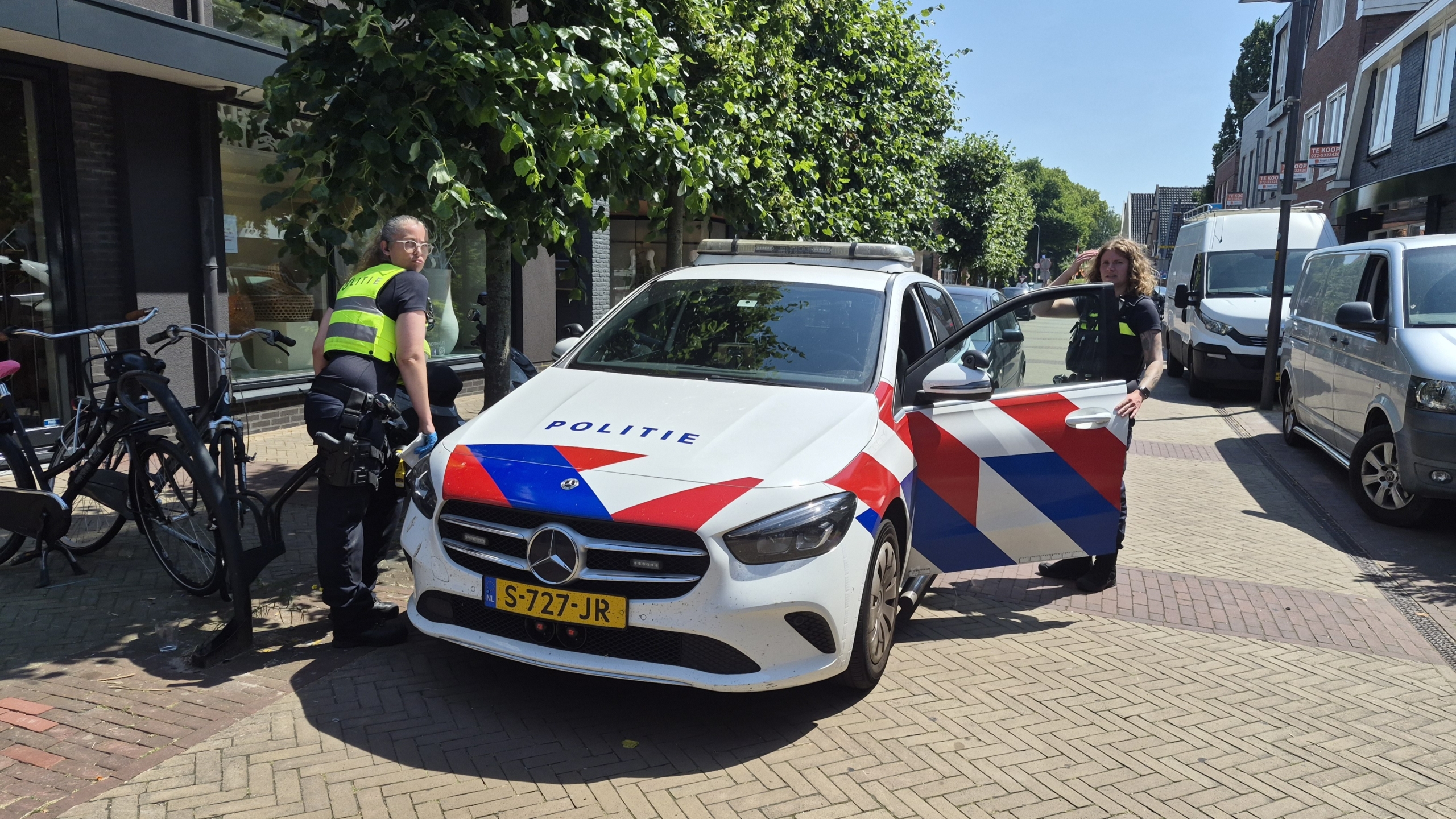 Twee politieagenten staan naast een politieauto op een straat met bomen en geparkeerde auto's.