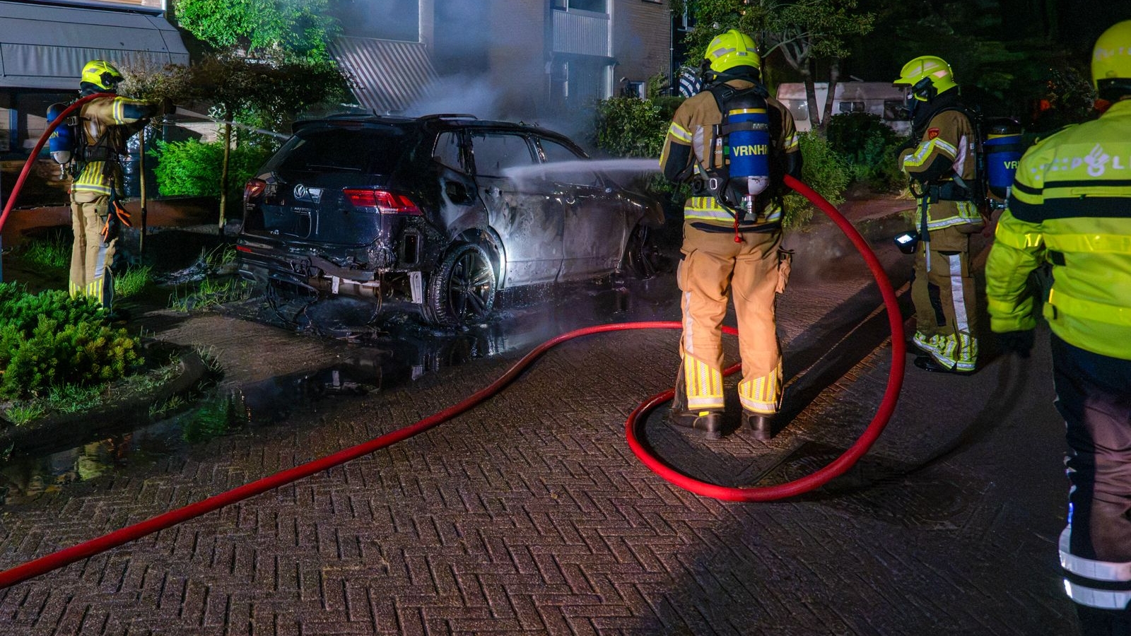 Brandweerlieden blussen een brandende auto op een straat 's nachts, omringd door rook en water.