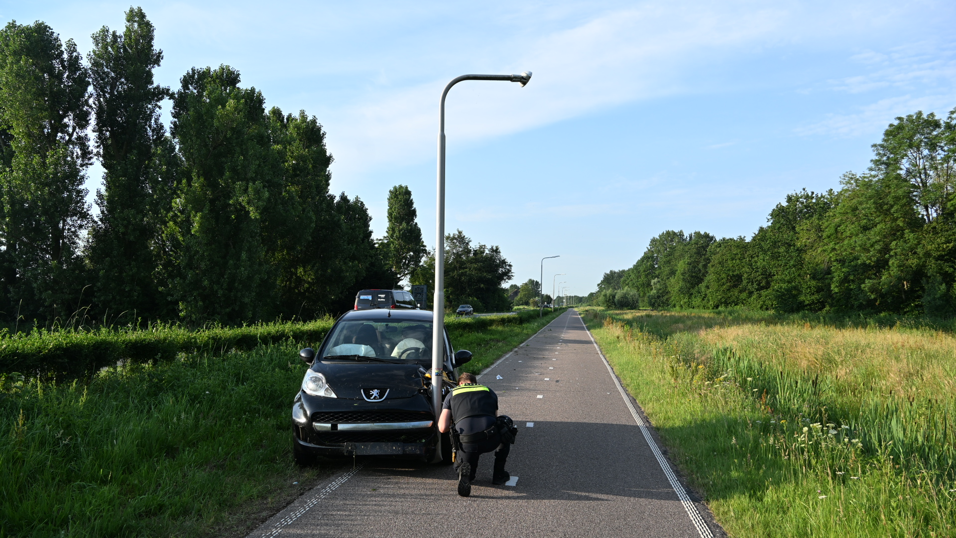 Auto gebotst tegen lantaarnpaal op rustige weg, met agent die schade inspecteert.