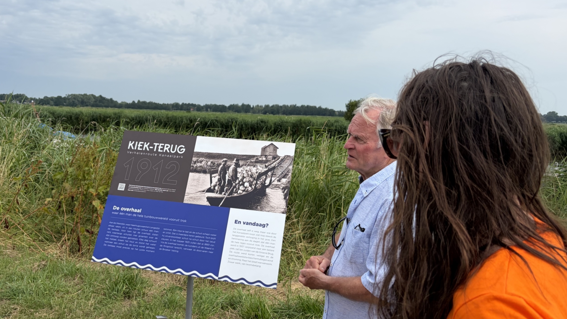 Een man en vrouw kijken naar een informatiebord met de titel "Kiek-Terug 1912" in een groen landschap.