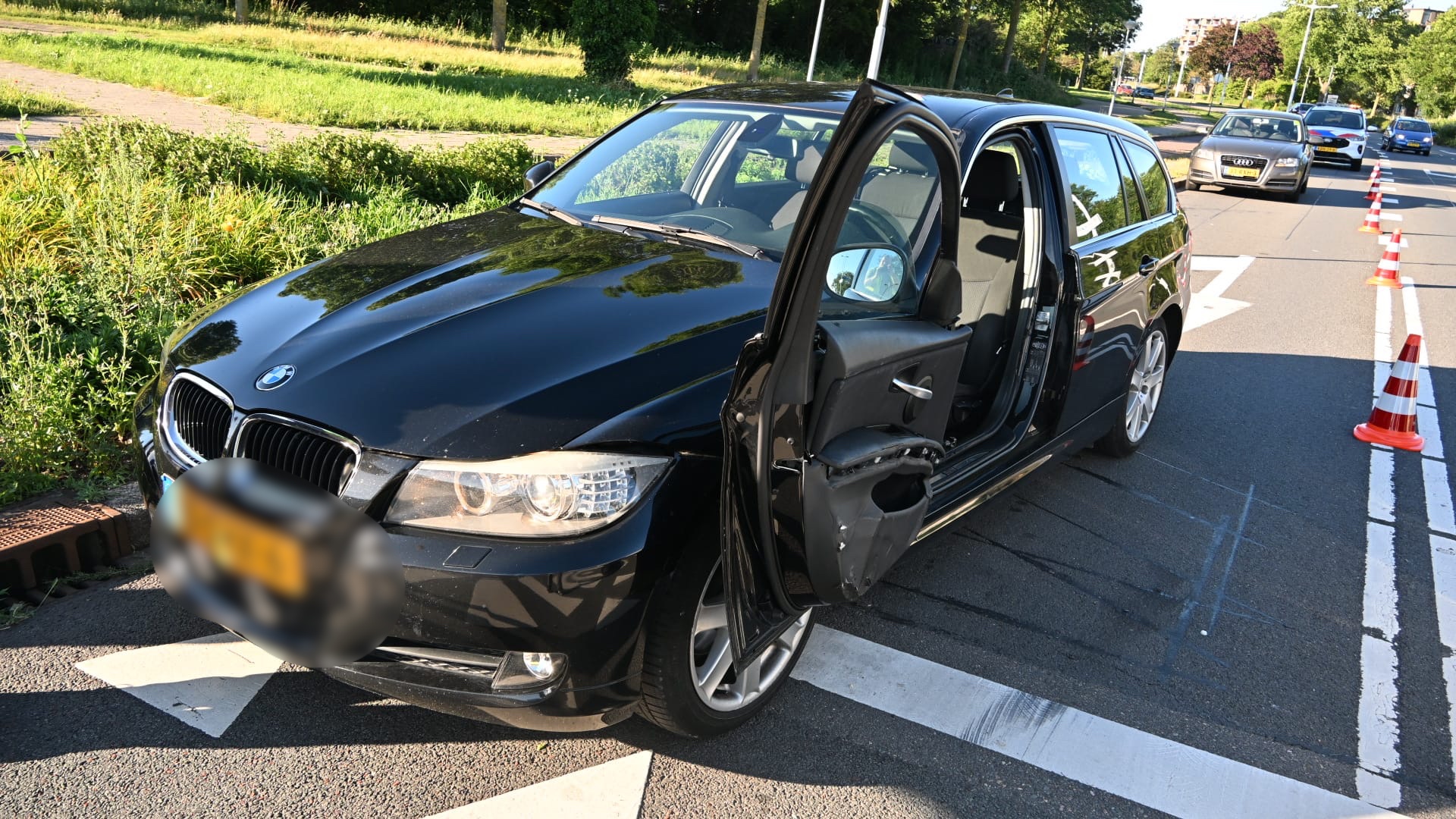 Zwarte auto met open achterdeur staat op een verkeersweg met kegels; andere auto's staan in de rij.