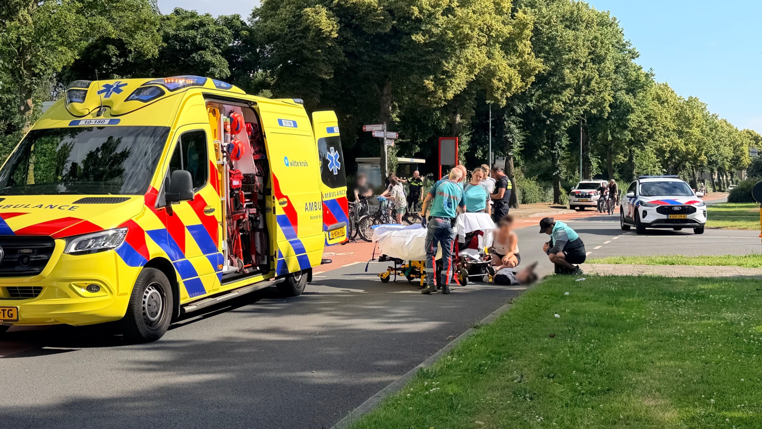 Ambulancepersoneel biedt hulp aan een persoon op de grond naast een ambulance en politieauto op een weg omringd door groen.