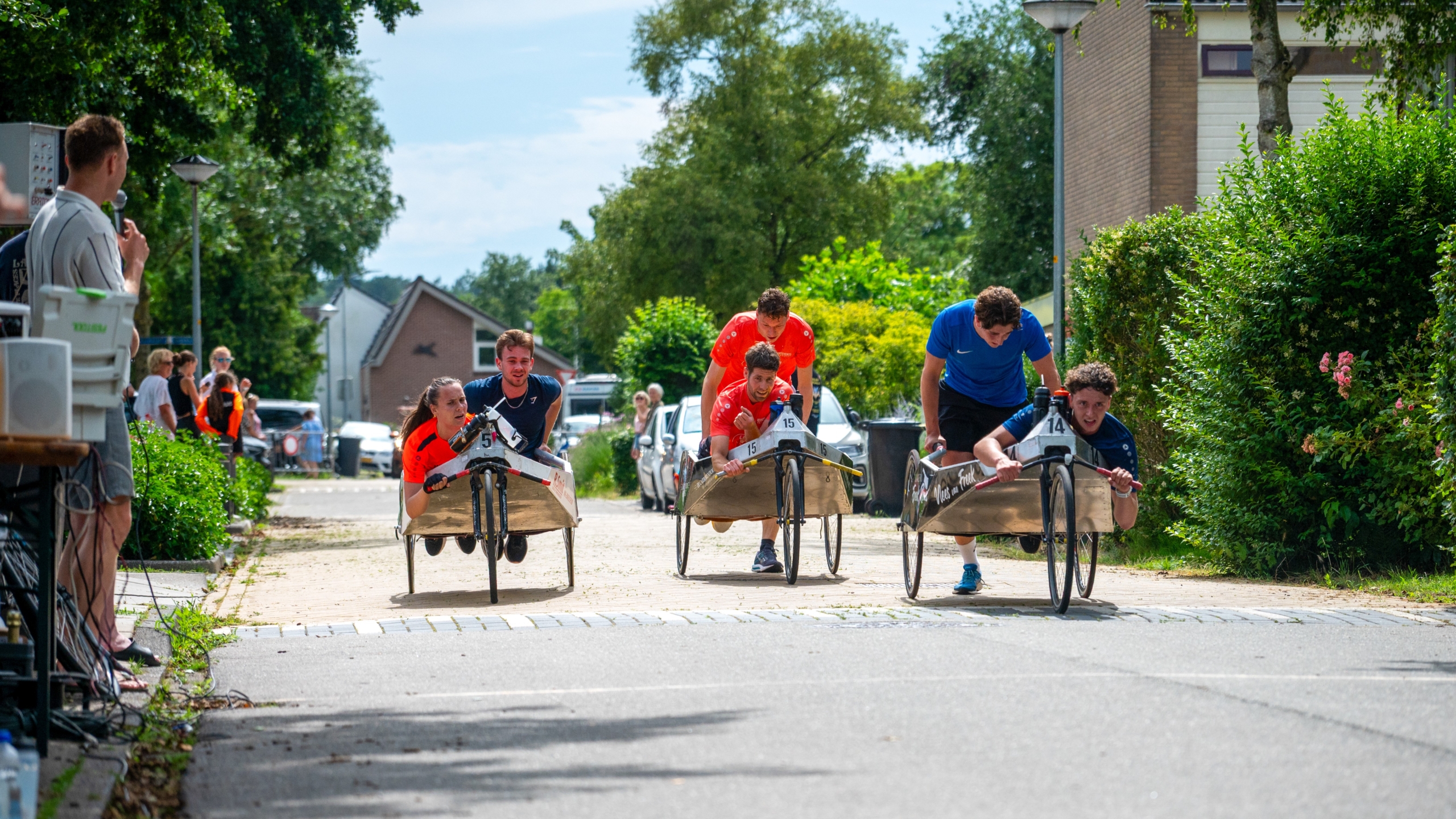 Mensen racen in zelfgemaakte karren over een straat tijdens een evenement met toeschouwers langs de kant.