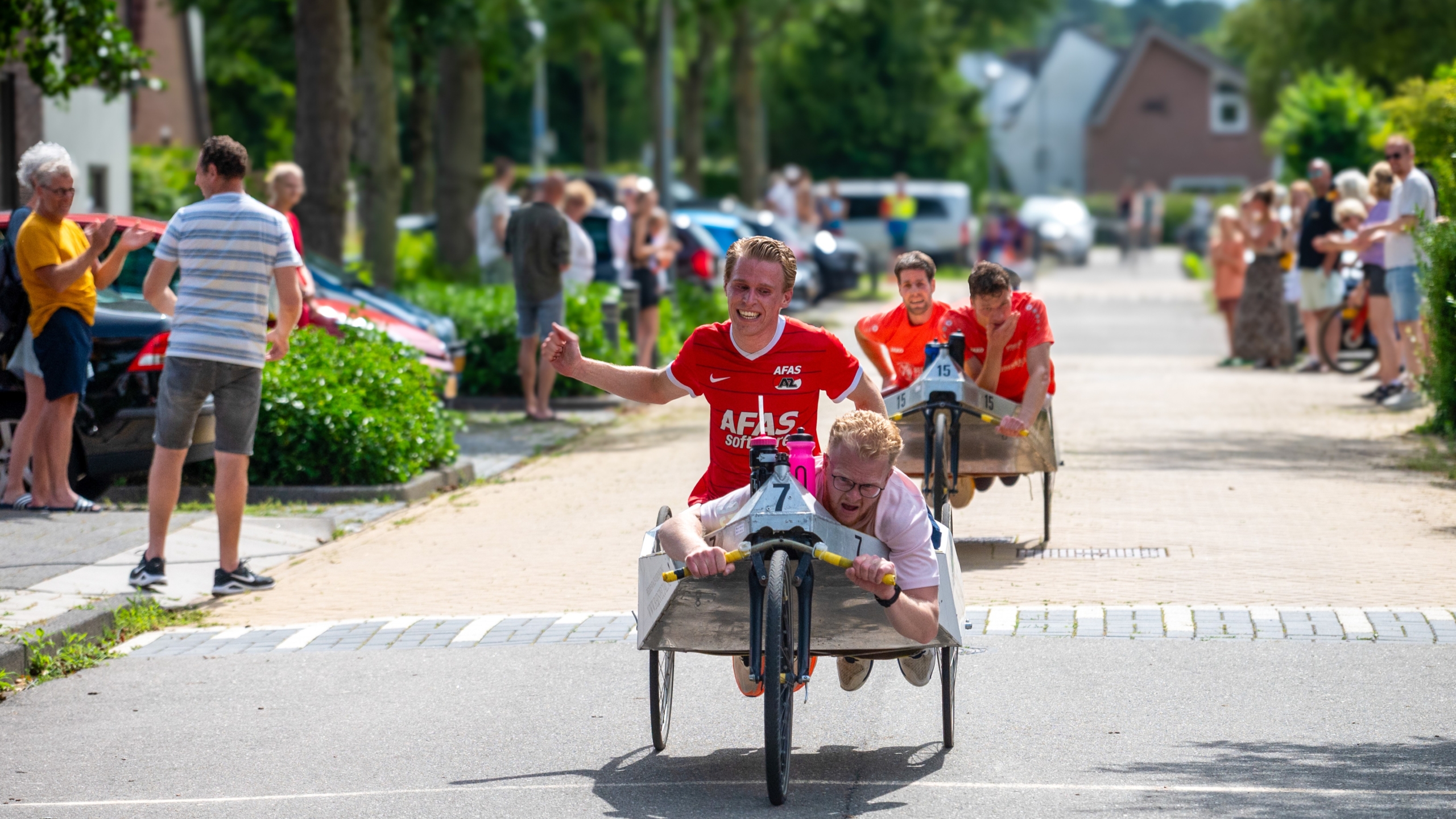 Mensen nemen deel aan een karretjesrace op een zonnige woonstraat, omringd door toeschouwers.