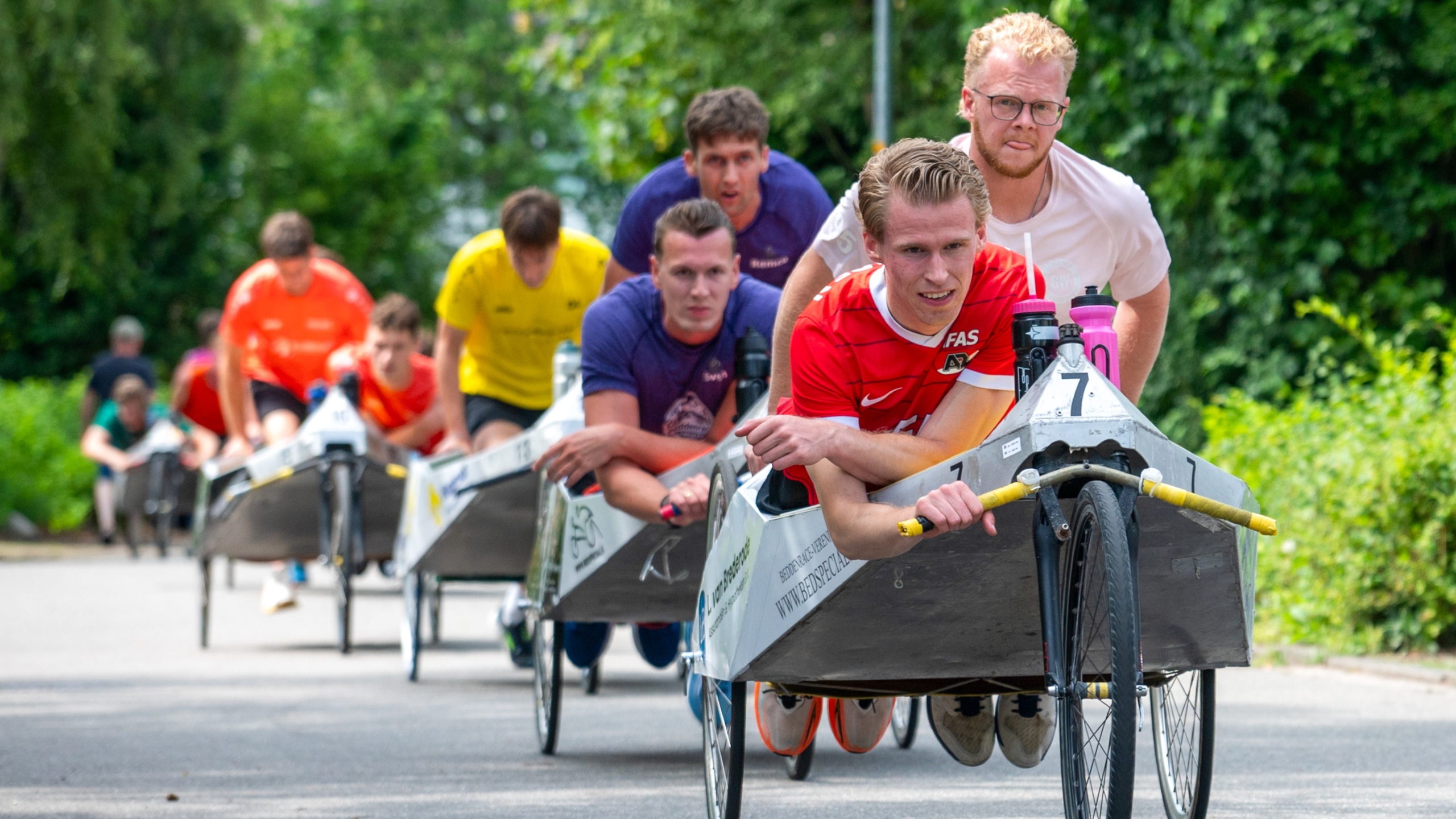 Een groep mensen racet in zeepkisten op een zonnige dag, gefocust en voorovergebogen terwijl ze aan het duwen zijn.