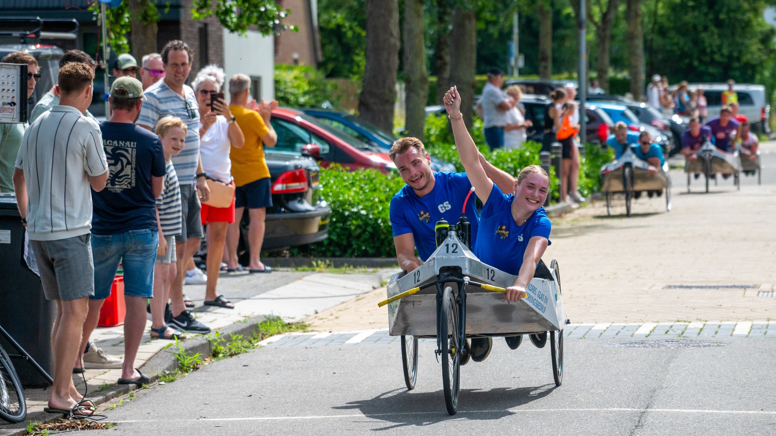 Twee mensen in blauwe shirts racen in een kar tijdens een evenement, omringd door toeschouwers langs de weg.