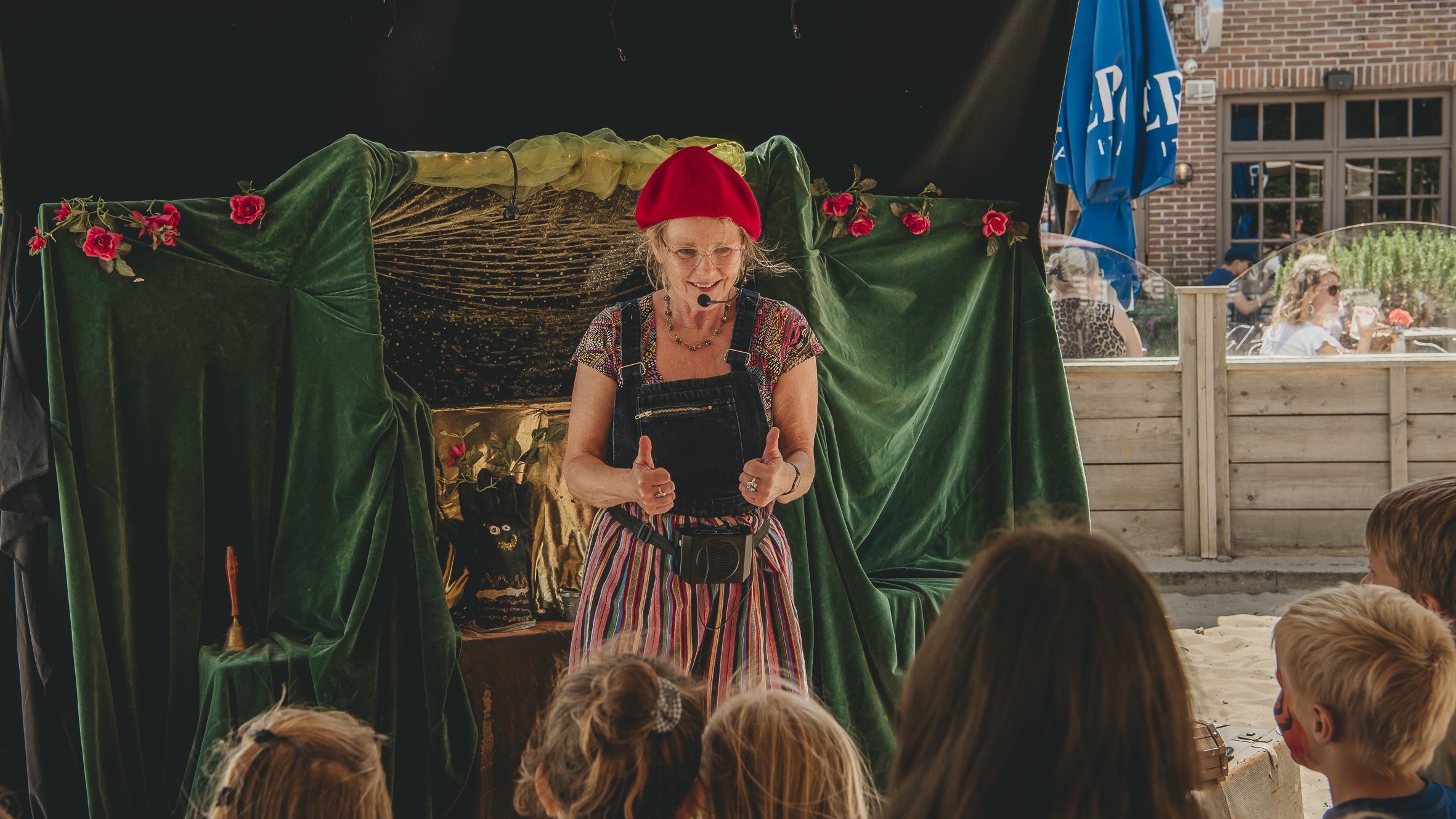 Vrouw met een rode baret geeft een voorstelling voor kinderen, omringd door fluwelen gordijnen en bloemen.